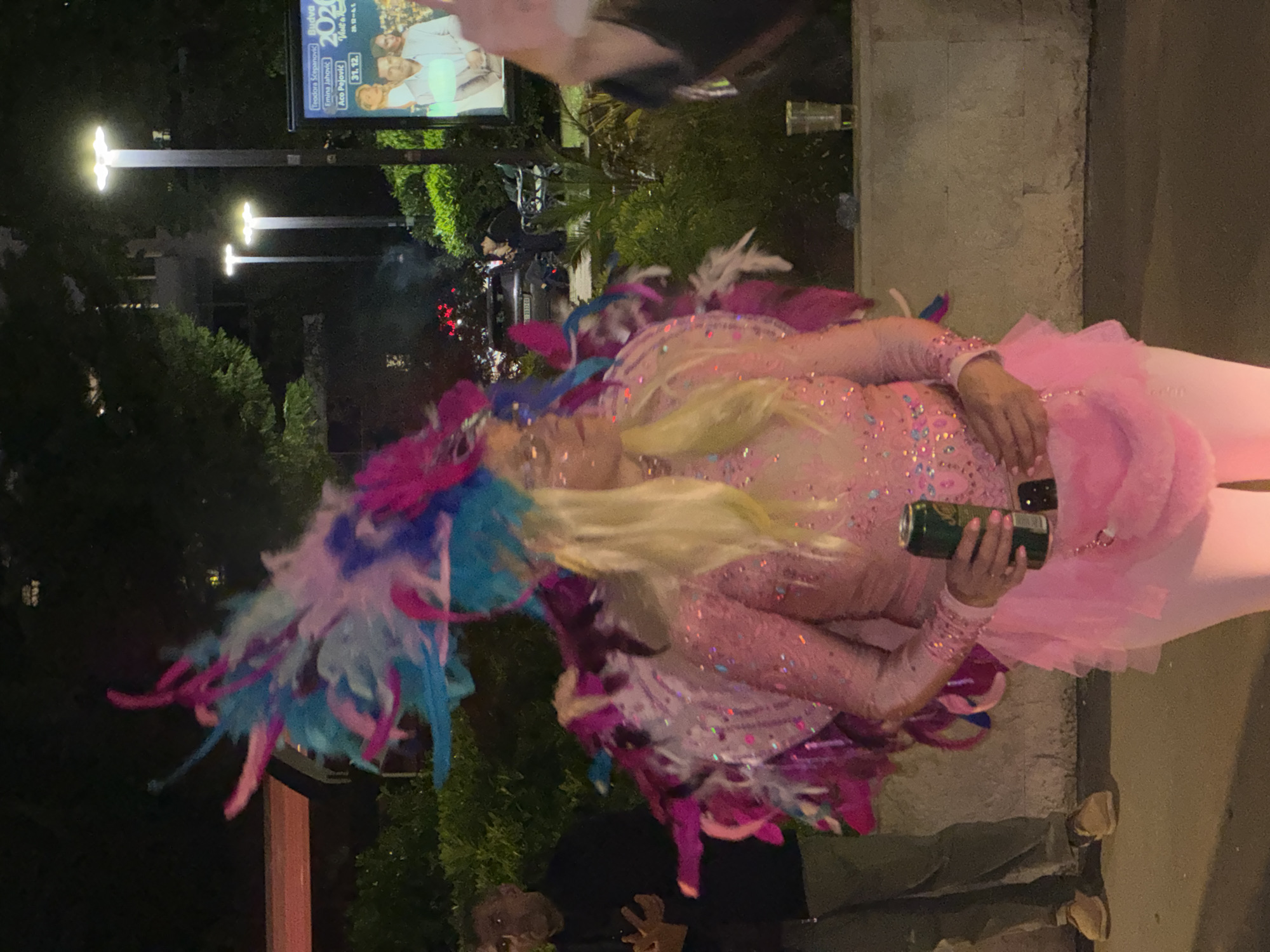 A color outdoor shot taken at night shows a person wearing a pink sequined costume with a colorful feather headdress, holding a beverage can and a mobile phone. The background shows streetlights and a billboard advertising events in Budva, Montenegro. Additional people and vehicles are visible in the background, along with greenery. A section of the pavement is in the foreground.