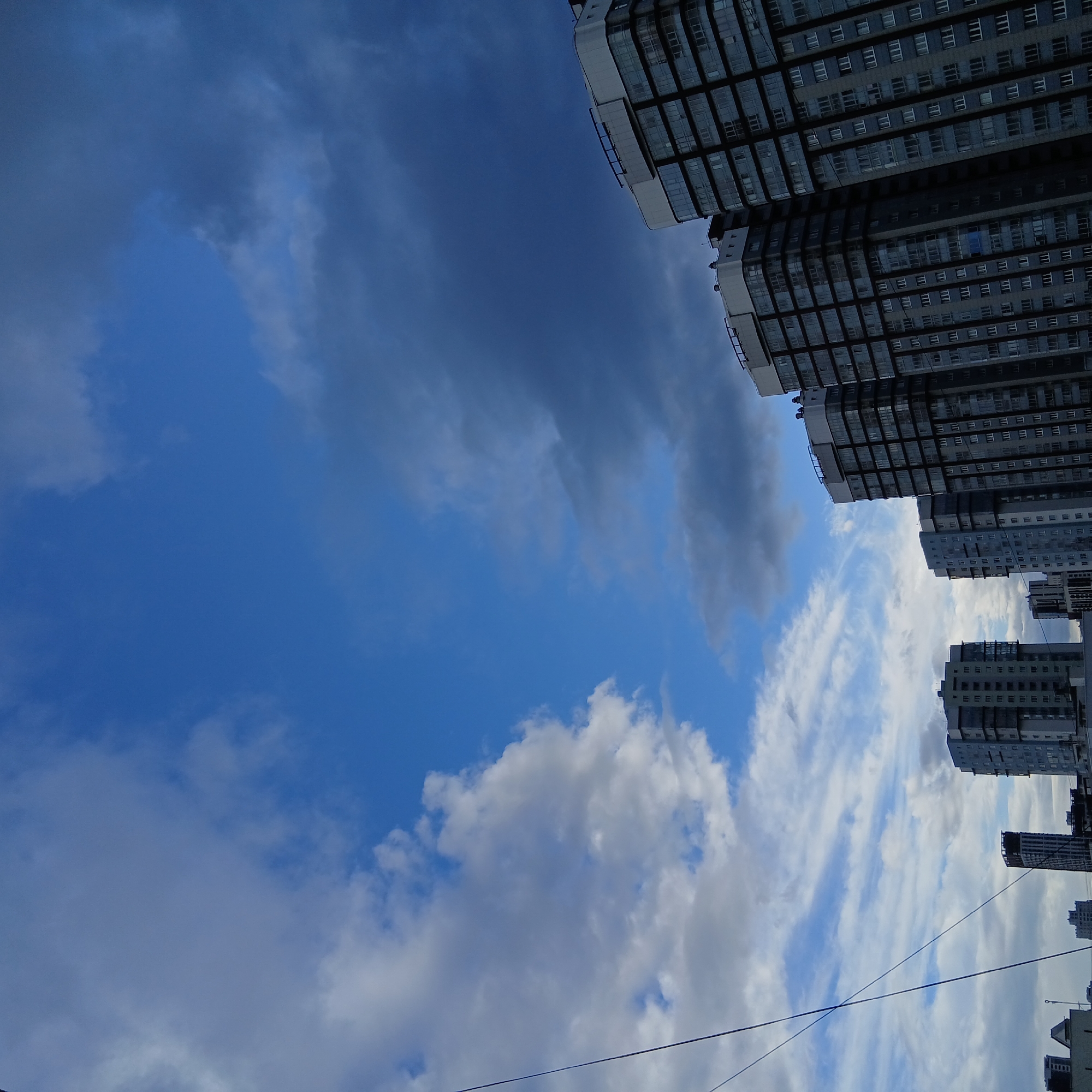 The image presents a dynamic upward view of a modern urban landscape under a partly cloudy sky. The perspective is tilted, with multiple high-rise buildings framing the sky from the right side of the frame, appearing to lean into the composition. These buildings are characterized by their grid-like facades of numerous windows and dark panels, suggesting contemporary glass and concrete architecture.
The sky dominates the central and left portions of the image, displaying a mix of deep blue patches, scattered white cumulus clouds, and larger, darker grey clouds that suggest either overcast conditions or impending weather. The contrast between the bright blue and the dark clouds is striking. Towards the bottom and right, faint power lines are visible, stretching across the sky, adding another element of urban infrastructure to the scene.
The time of day appears to be daytime, with ample natural light illuminating the clouds and building facades. No people or specific activities are visible within the frame. There is no discernible text. The scene depicts a typical cityscape from a worm's-eye view, characteristic of an urban environment like Yekaterinburg, Russia, with its blend of tall structures and expansive skies.