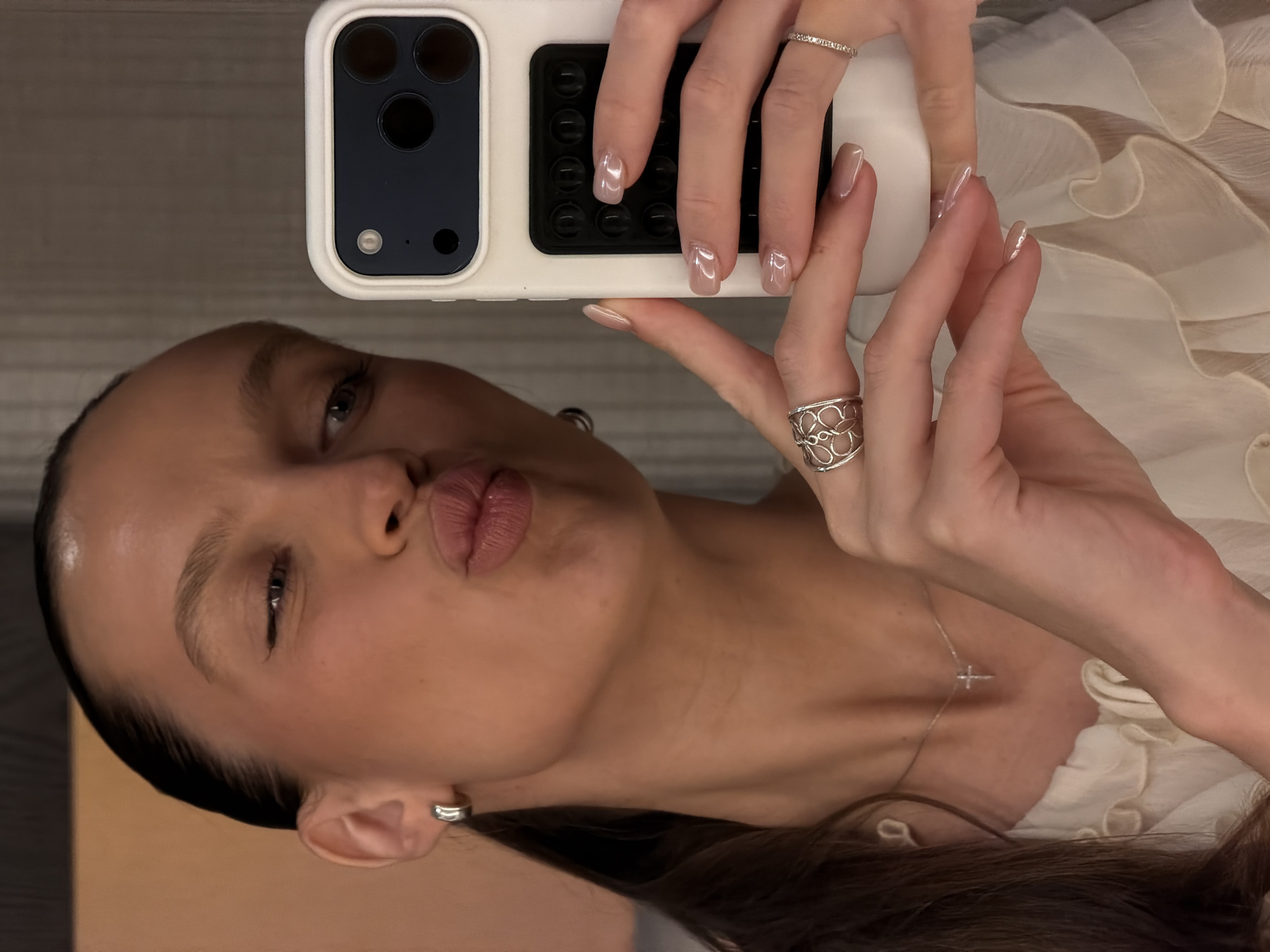 A woman with dark hair pulled back in a ponytail takes a selfie in a mirror. She is wearing a white ruffled blouse, silver hoop earrings, a silver necklace with a cross pendant, and two rings on her right hand. One ring is a silver band with small stones, and the other is a larger silver ring with a floral design. Her nails are light pink and appear to have a pearlescent finish. She is puckering her lips for the camera. The background is a blurred interior. There is no indication of Budva, Montenegro in the image.
