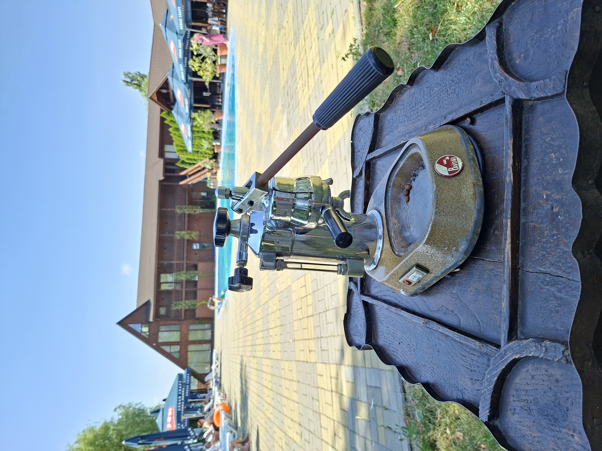 A vintage Pavoni espresso machine, featuring a chrome lever and a brass boiler, rests on a dark, weathered wooden table. The machine has a prominent red power switch and a glass water level indicator. Several coffee beans are visible in the drip tray. The table is positioned outdoors on a sunny day, with a swimming pool and a large wooden building in the background. The building appears to be a resort or hotel, with balconies and several windows. Umbrellas and lounge chairs are set up around the pool area, suggesting a recreational setting. In the distance, people can be seen relaxing by the pool and under the umbrellas. The overall scene depicts a serene outdoor setting, possibly a cafe or a hotel courtyard in Costești, Moldova.