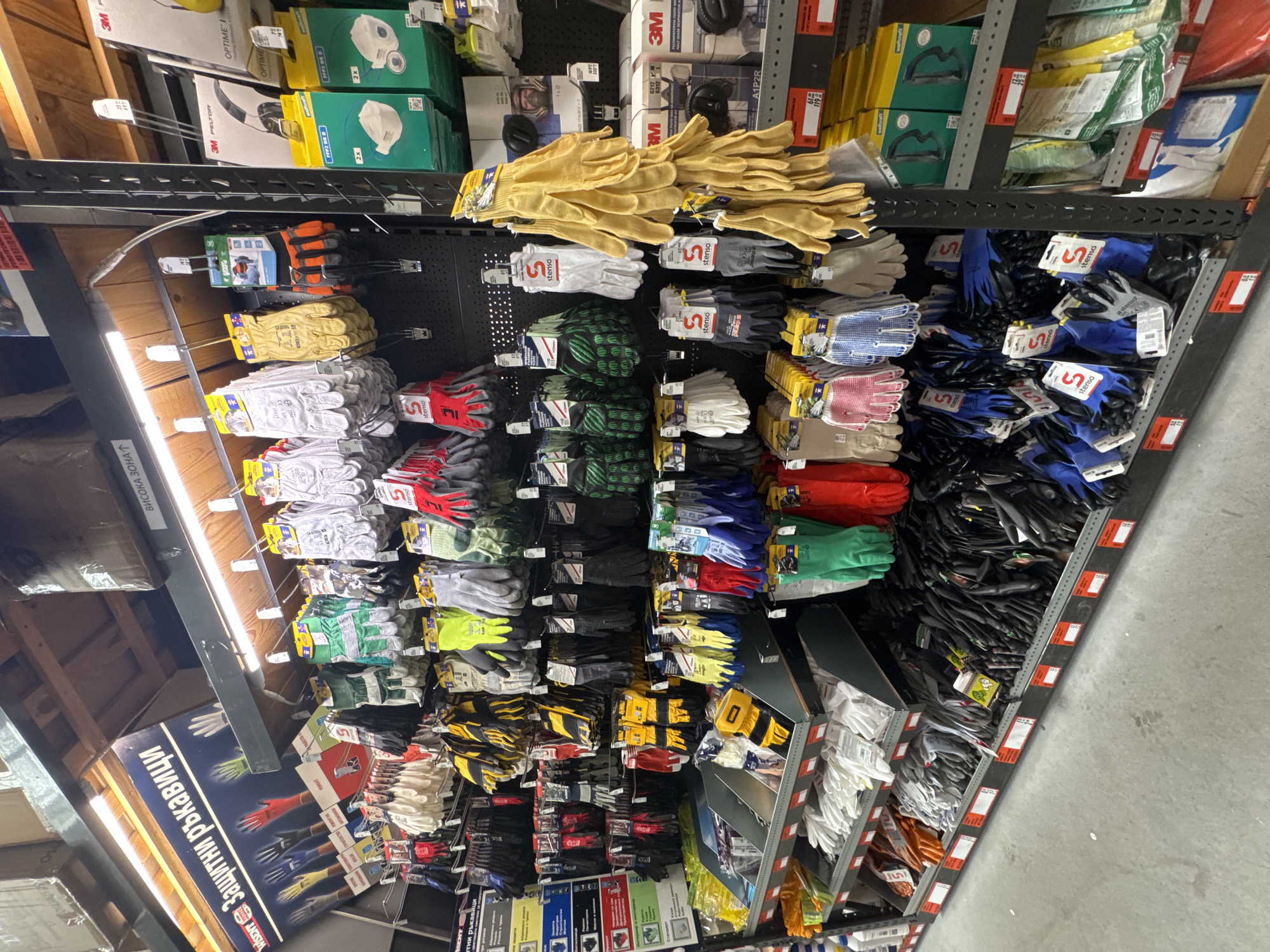 A low-angle, color photograph shows a store display of work gloves and 3M safety equipment. The shelves are stocked with gloves of various materials and colors, including yellow leather, white cotton, and blue nitrile. Some gloves bear the brand name "Stenso." Above the gloves are shelves containing 3M Peltor Optime I hearing protection and 3M respirators. A sign with the text "ЗАЩИТНИ РЪКАВИЦИ" (Protective Gloves) and "ВИСОКА ЗОНА" (High Zone) is visible at the bottom left. This suggests the image was taken in a store in Sofia, Bulgaria.