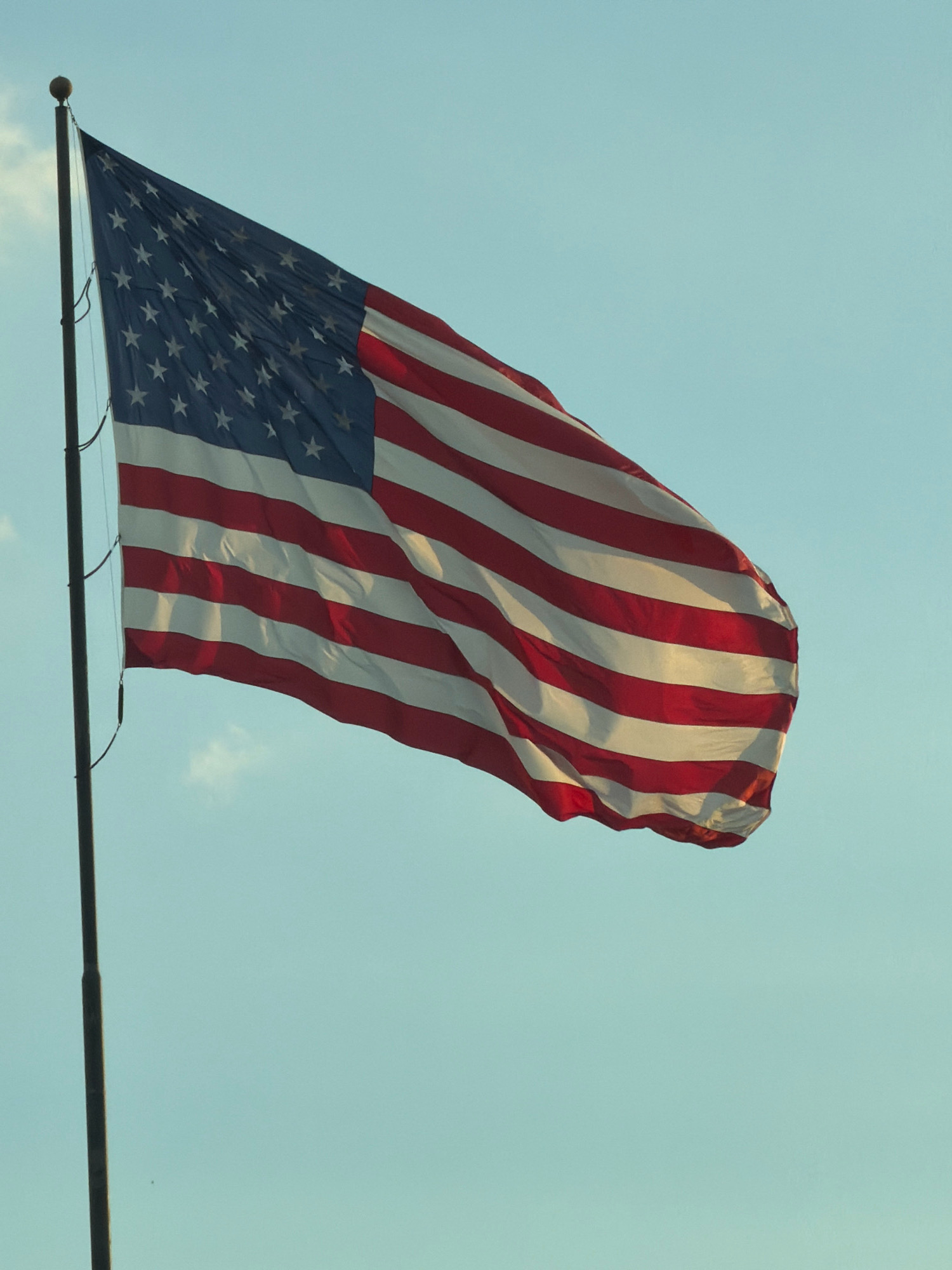 An American flag flies on a flagpole against a clear, pale blue sky. The flag is predominantly red and white striped with a blue canton containing white stars. The flag is billowing in the wind. The flagpole is dark and appears to be made of metal, with a spherical finial at the top. There are no people visible in the image. The scene appears to be outdoors, possibly in a park or open area. The specific location in San Marcos, United States of America is not identifiable from the image alone.