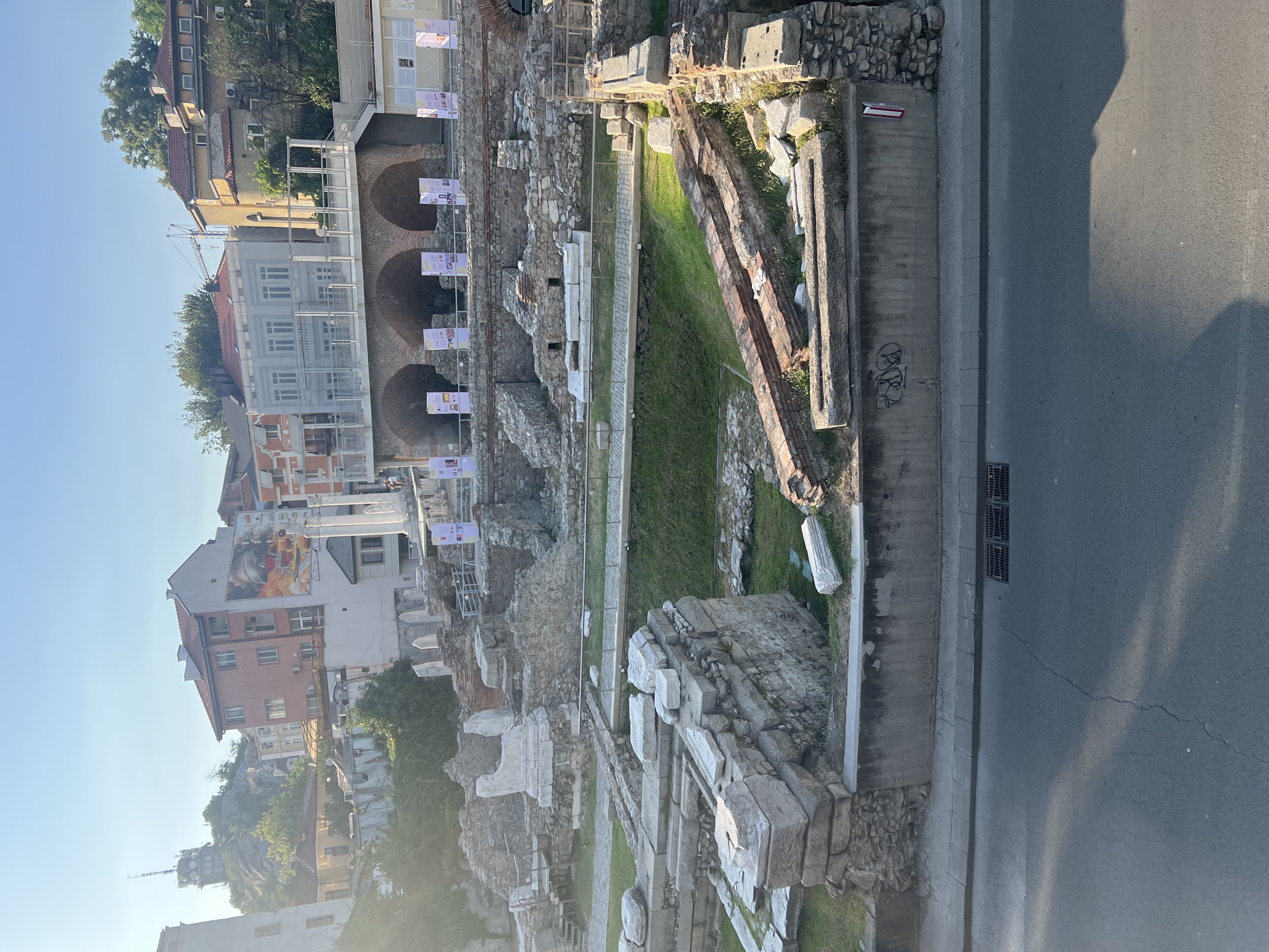 The image captures a view of the Roman Stadium of Philippopolis in Plovdiv, Bulgaria, looking north. In the foreground, a section of an asphalt road with a visible drain grate and some shadows is present. Above the road, a concrete wall with graffiti is visible, topped with ancient stone ruins, including large blocks and what appears to be a sarcophagus-like structure.
The middle ground showcases the excavated remains of the Roman stadium, characterized by stone foundations, walls, and fragments of columns. Green grass covers some of the excavated areas, and pathways wind through the ruins. Banners displaying information are positioned in front of the stadium's arched structures.
In the background, several buildings of varying architectural styles and heights are visible. The most prominent is a tall, modern building with a large advertisement on its side. Further in the distance, a hill is topped with a structure, possibly a communication tower, against a clear blue sky. Trees are scattered throughout the urban landscape. The overall scene depicts a blend of ancient history and modern urban life.