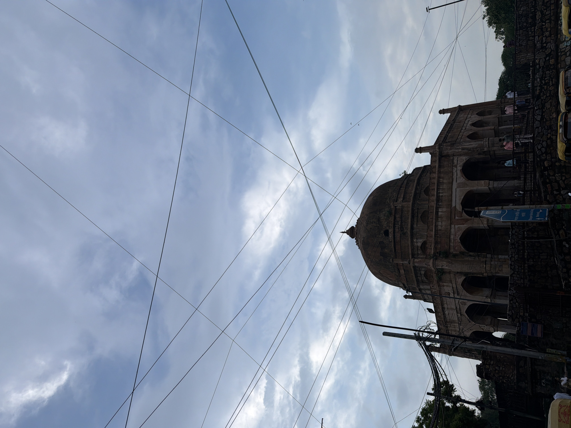 This image captures a historical dome structure in Delhi, India, under a cloudy sky. The perspective is from a low angle, looking upwards, with numerous power lines crisscrossing the frame, creating a dense, almost chaotic pattern against the sky. The dome itself appears to be made of weathered stone, with arched openings visible on its lower sections. The surrounding area shows a stone wall and some greenery, suggesting an older, possibly historical site.
The time of day appears to be daytime, likely overcast due to the cloudy sky. The weather seems humid and possibly warm, typical of Delhi's climate. A few people are visible in the image, standing near the structure or walking by, indicating it is a public space. The presence of yellow auto-rickshaws on the right side of the image provides a modern contrast to the ancient architecture and reinforces the location as India.
There is some visible text on a blue sign in the lower right quadrant of the image, which appears to be in Hindi. However, the text is blurry and not legible enough to decipher its specific content. The overall mood of the image is somewhat stark, with the interplay of the old architecture and the modern, intrusive power lines creating a sense of a place caught between eras.