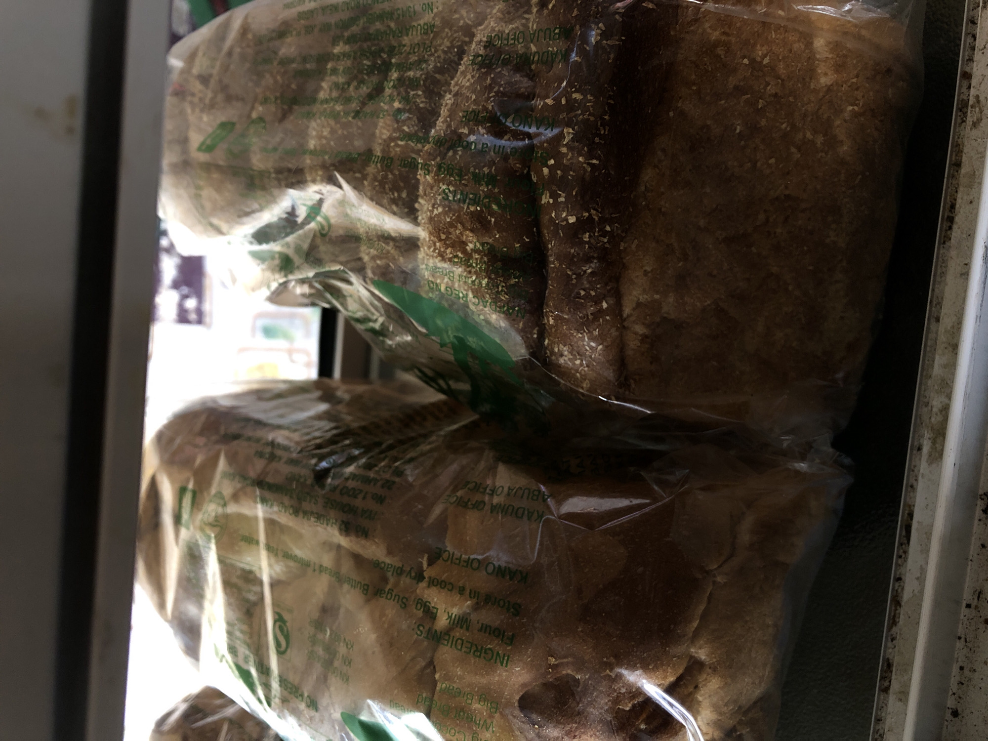 The image displays two loaves of bread in plastic packaging, likely on a shelf in a shop or bakery in Jos, Nigeria. The bread is a brown, whole wheat variety, with the crust visibly textured. The plastic packaging features text that includes "INGREDIENTS:", "Flour, Milk, Egg, Sugar, Butter/Bread", and storage instructions. There are also company details like "KANO OFFICE", "KADUNA OFFICE", and "ABUJA OFFICE", suggesting a multi-city operation. The lighting is somewhat dim, indicating an indoor setting. The focus is on the bread loaves, with the background slightly blurred. The bottom of the image shows a dirty, white ledge or surface.