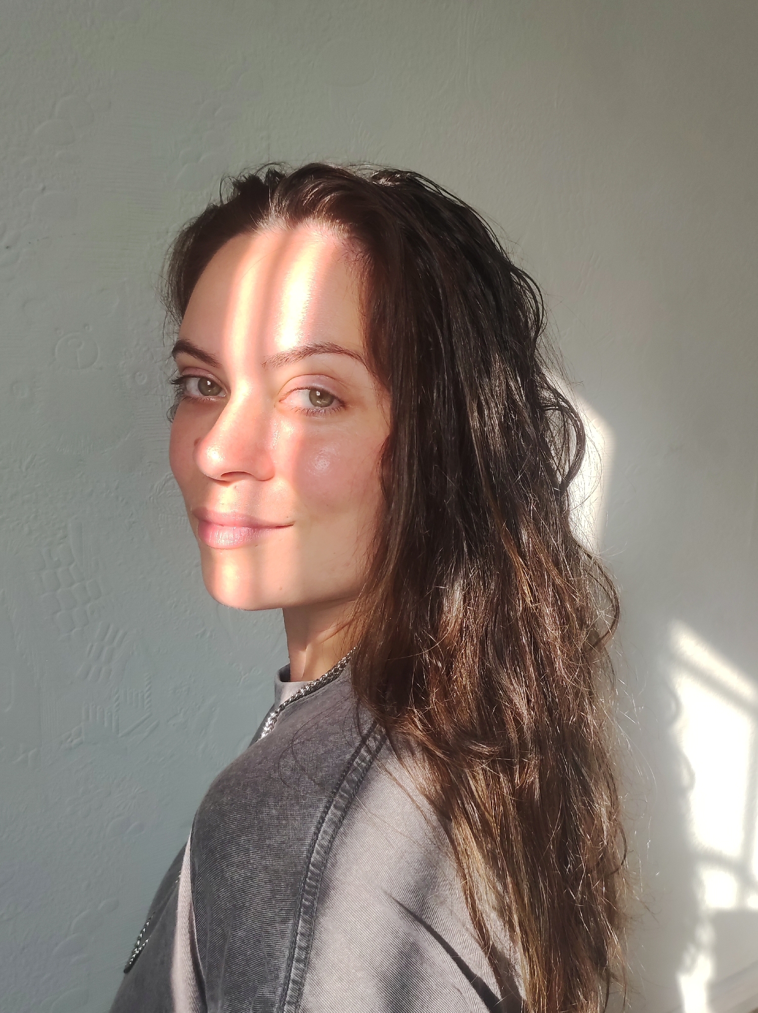 The image is a medium close-up shot of a woman with long, dark brown hair. She is looking over her shoulder at the camera, with her head tilted slightly. Her skin appears to be glowing, possibly due to the warm sunlight that is casting striped shadows across her face and the white wall behind her. She is wearing a grey top with a chain detail around the collar. The wall has a textured pattern. The scene is indoors, possibly in a room with a window allowing light to stream in. There is no specific information to identify this as being in Chaikovsky, Russia.
