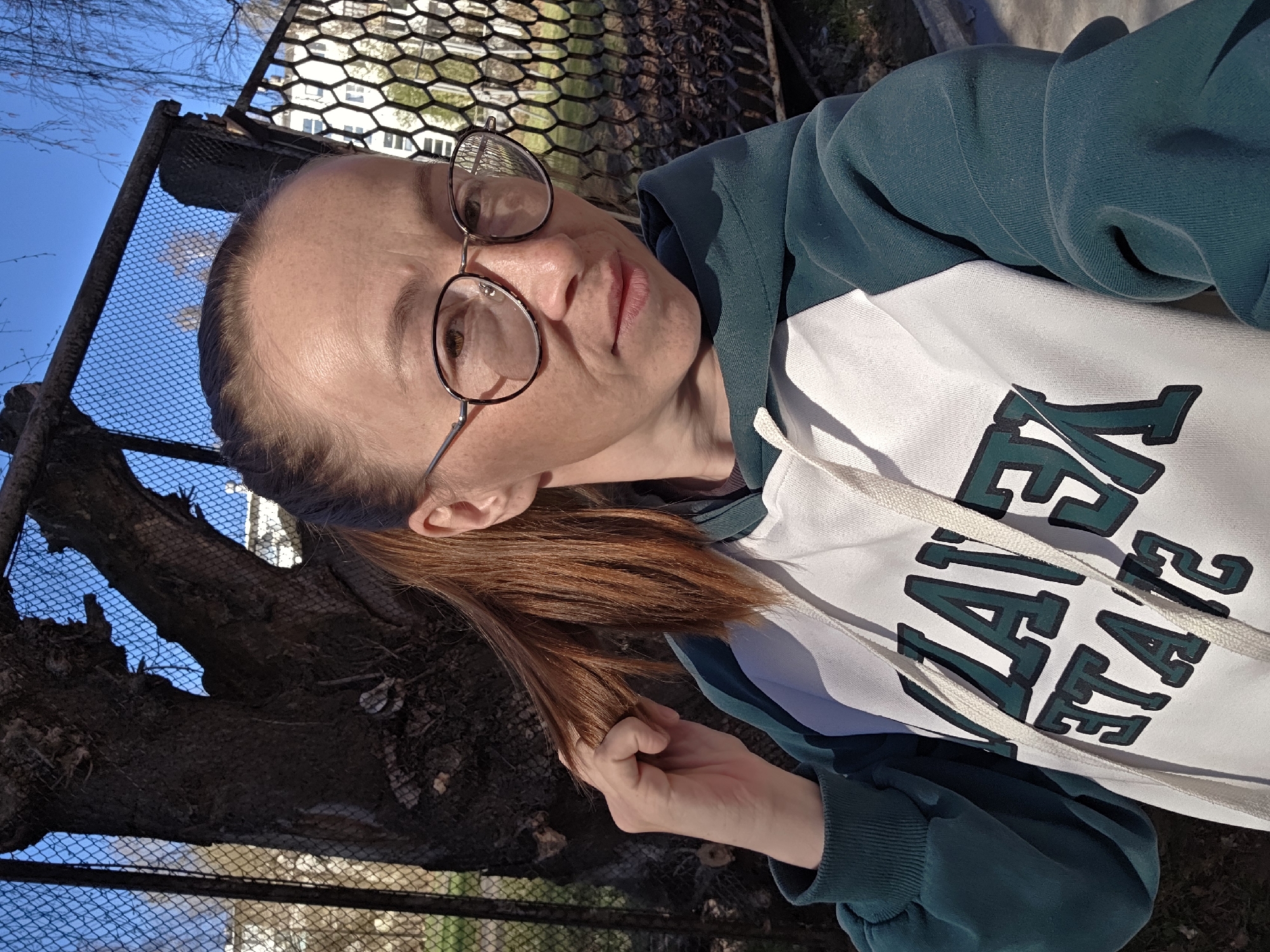 A woman is seen in a close-up, selfie-style shot. She is wearing glasses and a two-tone hoodie, with green sleeves and a white chest. The hoodie features dark green lettering. She is holding a strand of her reddish-brown hair. The background shows a chain-link fence and a tree branch, with a blue sky visible through the fence. The location appears to be an outdoor setting. Based on the request, this is in the city of Chaikovsky, Russia.