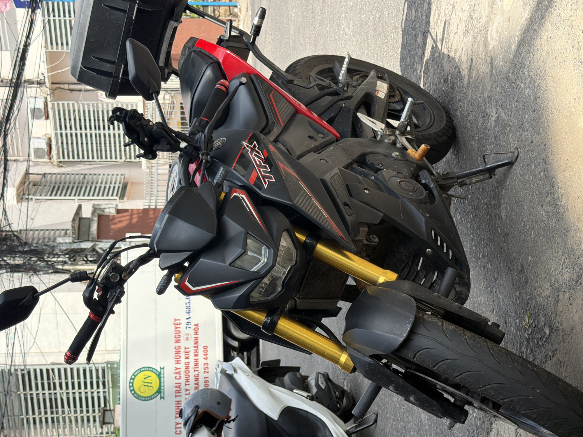 This image depicts a black and red Yamaha TFX motorcycle parked on an asphalt street, likely in Nha Trang, Vietnam, given the context. The motorcycle is the primary subject and is viewed from a low angle, emphasizing its front and side. It has gold-colored front forks, a black seat, and a black storage box mounted on the rear. A white scooter is partially visible to the left of the motorcycle. In the background, buildings with striped window coverings and air conditioning units are visible, along with electrical wires overhead. A white truck with a circular green logo featuring the letters "HN" and text that appears to be in Vietnamese is parked behind the scooter. The ground shows signs of dust and debris, consistent with an urban outdoor environment. No people are present in the image.