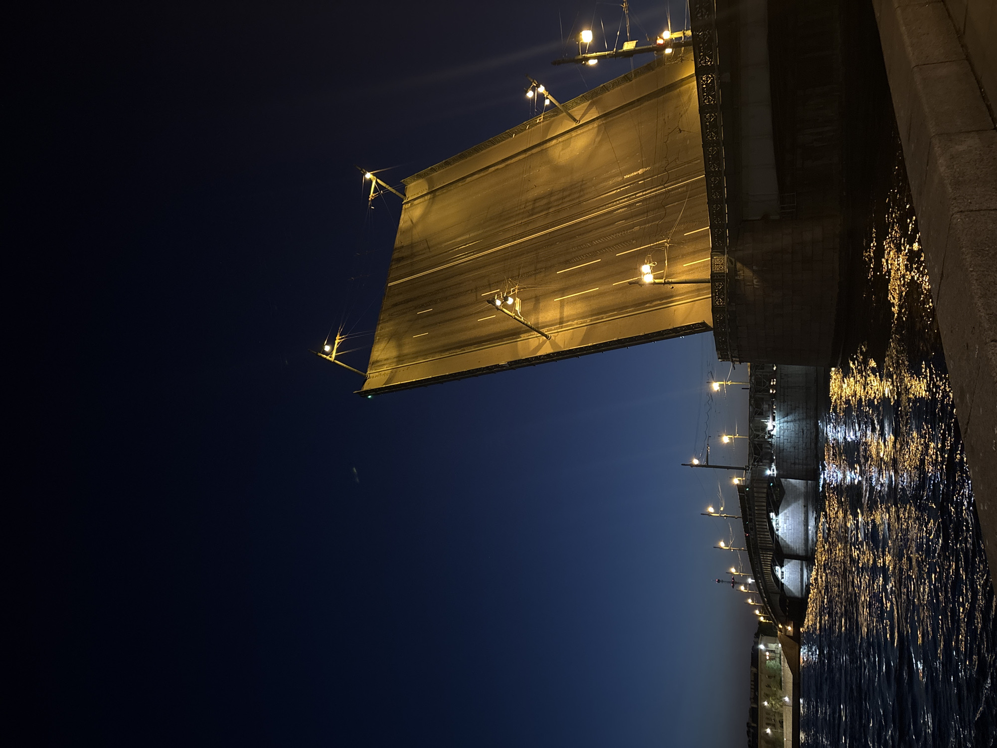 The image depicts a bridge over water at night. The bridge is illuminated by streetlights and other sources, casting reflections on the water's surface. The sky is dark, transitioning from a deep blue to black. The water is rippled and reflects the bridge lights. The bridge appears to be a drawbridge based on the lifted section on the left side of the frame. The location is the city of Unknown, Unknown.