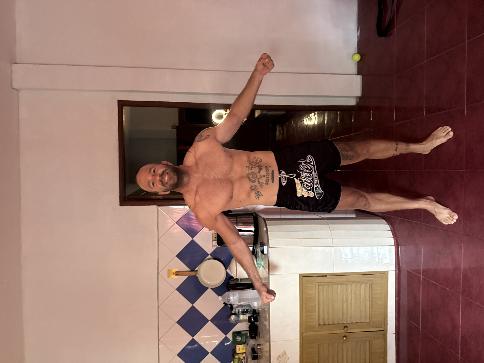 A bald, shirtless man with tattoos stands in a kitchen in Ko Samui District, Thailand, with his arms outstretched. He is wearing black Muay Thai shorts. The kitchen has blue and white checkered tiles on the wall, a wooden cabinet, and a doorway leading to another room. A water bottle and other items are on the counter. The floor is tiled in a dark red. A yellow tennis ball is on the floor to the right of the man.