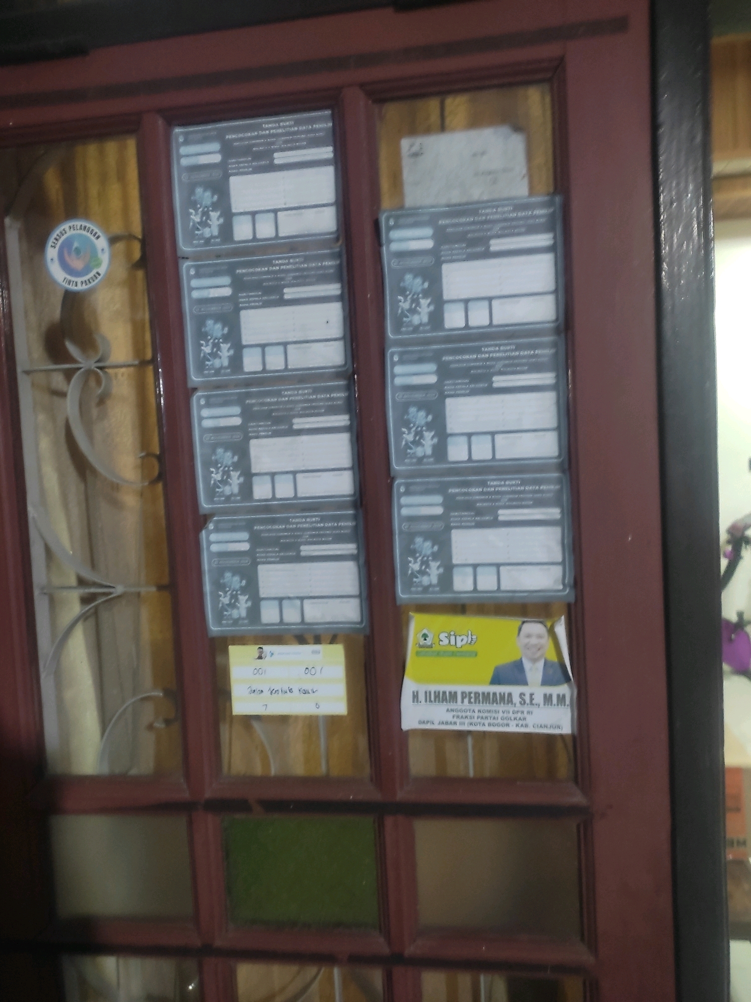 The image shows a closed wooden door with glass panes. Several documents are affixed to the door with tape. The documents appear to be official forms with text in Indonesian. A sticker with a circular logo containing a globe and text that appears to say "SERBU PELANANAN" and "TINTA PASAR" is visible on the left-hand side of the door. On the right side of the door, a flyer-like document features a portrait of a man in a suit, identified as "H. ILHAM PERMANA, S.E., M.M. ANGGOTA KOMISI VII DPR RI FRAKSI PARTAI GOLKAR DAPIL JAABAR III (KOTA BOGOR - KAB. CIANJUR)". A yellow sticky note is also attached to the door with handwritten text. The environment suggests an indoor setting, possibly a building entrance, given the presence of official documents and a political flyer. The overall appearance of the door and the way the documents are displayed suggest this location is in Bogor, Indonesia, due to the Indonesian language on the documents and the political figure's affiliation with a specific electoral district in Indonesia.
