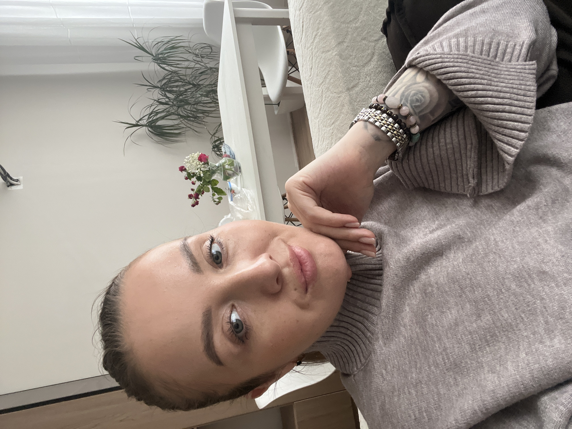 The image shows a private residence, specifically an interior space resembling a living room or dining area, in an unknown city. A woman, appearing to be in her 20s or 30s, occupies the foreground, facing the camera directly with a slight smile. She has light-colored eyes and her hair is pulled back. She is wearing a light grey, ribbed turtleneck sweater. Her right hand rests under her chin, while her left forearm, adorned with multiple bracelets of various materials (beads and metal), reveals a grey tattoo resembling a floral design. In the background, to the subject's right, a white rectangular table holds a clear glass vase containing red and white flowers, a white plastic bag, and a small bowl. Behind the table, a tall potted plant in a white planter and two white modern chairs with wooden legs are visible. The room features plain light-colored walls and a large window with sheer white curtains on the right. A section of a wooden cabinet is visible on the far left. A ceiling-mounted fixture is partially visible above the left side of the scene.