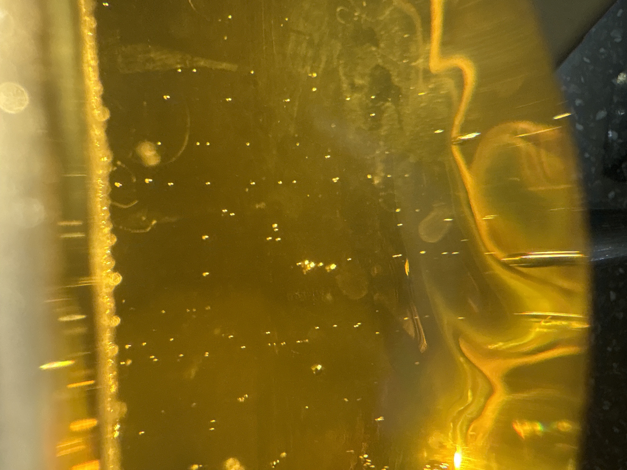 The image is a close-up, abstract shot of a viscous, golden-yellow liquid, likely oil or honey, with small bubbles suspended within it. The lighting creates bright highlights and streaks, emphasizing the liquid's texture and translucency. The bottom of the image shows a dark, textured surface, possibly a container or countertop. The image does not contain any information that can be used to determine its location in Georgiyevsk, Russia, or any specific context beyond the visual details of the liquid itself. There are no people, actions, or interactions depicted.
