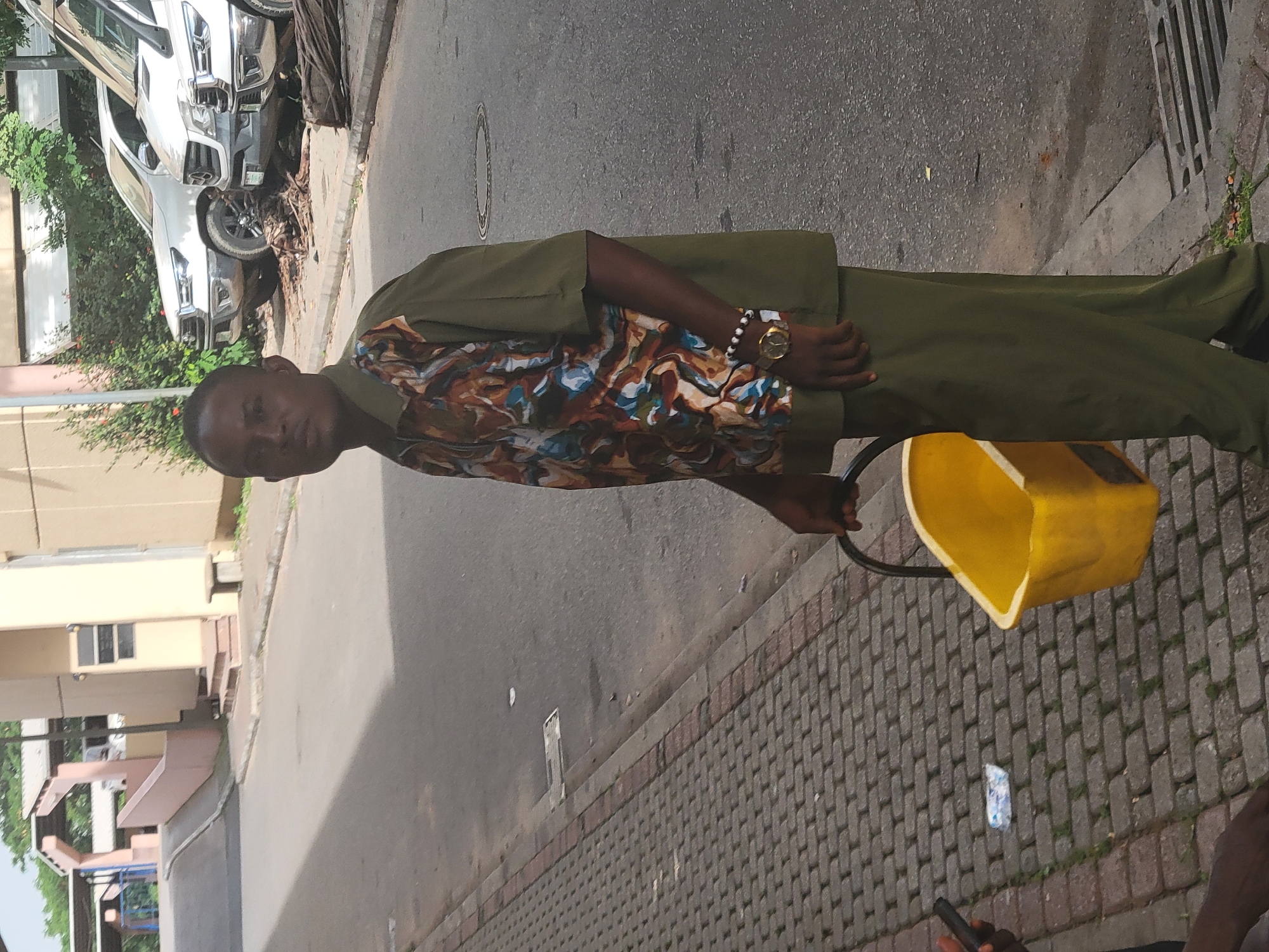 The image, oriented vertically, depicts a man standing on a paved sidewalk next to an asphalt street in an urban environment in Abuja, Nigeria. The man, with dark skin, is positioned slightly off-center and looks directly at the viewer with a neutral expression. He is dressed in an olive green long-sleeved shirt, which features a vibrant, abstract pattern of brown, blue, orange, and white hues across its front and sleeves. He also wears matching olive green trousers. On his left wrist, a gold-toned watch and a bracelet made of black and white beads are visible. His right hand grasps the black handle of a bright yellow, somewhat rectangular container or bucket, which rests on the gray paver sidewalk next to him.
In the background, across the street, several white pickup trucks or SUVs are parked along the curb, partially obscured by lush green foliage. The street itself is asphalt, and the sidewalk is constructed from grey interlocking pavers. Further in the background, light-colored buildings with multiple windows suggest a mix of residential or commercial structures. The lighting indicates it is daytime with clear weather conditions. A small piece of litter is visible on the sidewalk near the yellow container, but no discernible text can be read anywhere in the image.