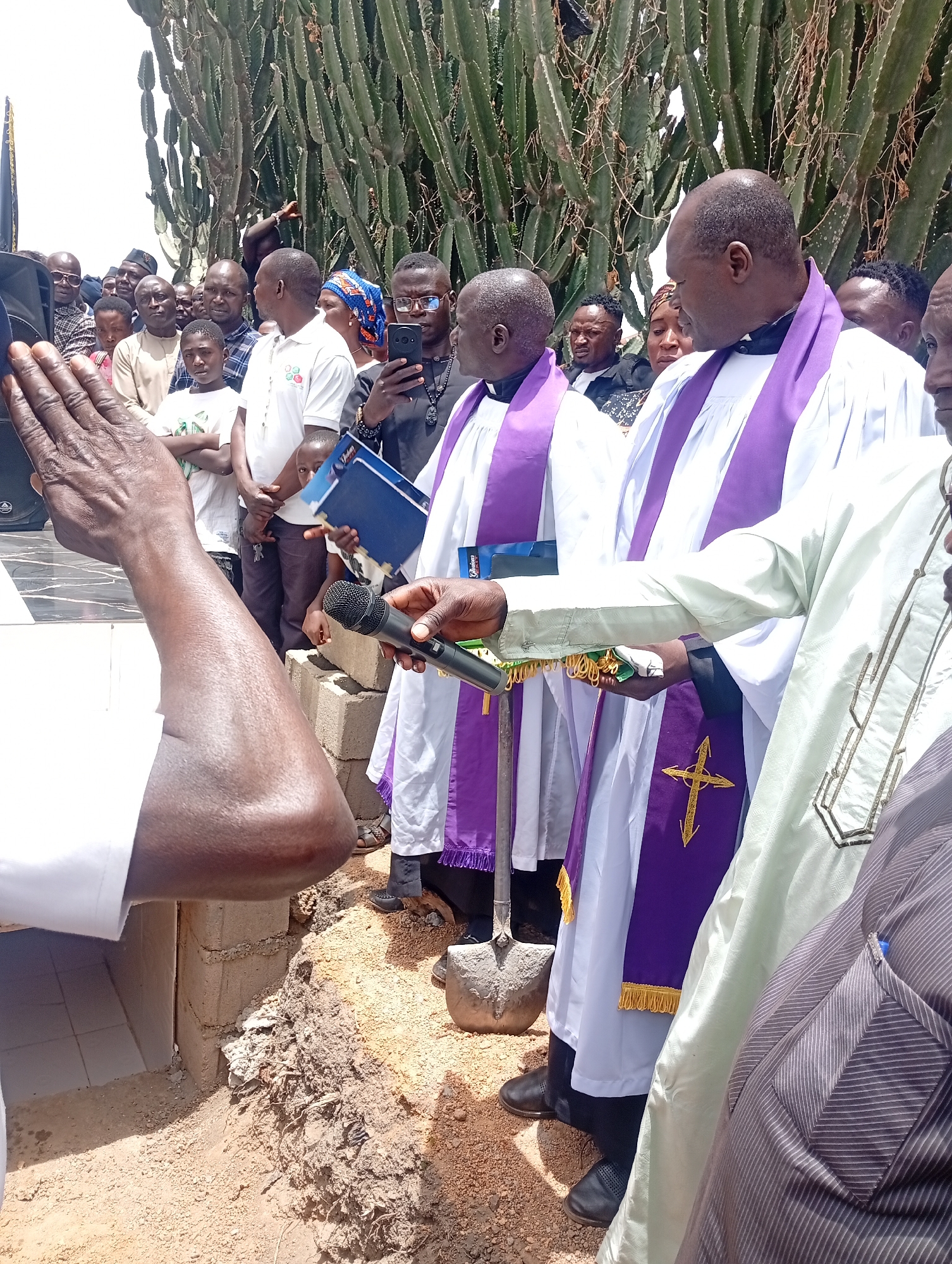 This image depicts an outdoor religious ceremony, likely a burial or a groundbreaking, taking place in Laduran Foron, Nigeria. The scene is set under bright daylight, with a large gathering of people, predominantly men, observing the proceedings.
In the foreground, a person's arm is raised with an open palm, seemingly in a gesture of prayer or blessing. Several Christian clergy members are central to the event, dressed in white robes and purple stoles, some embroidered with gold crosses. One clergyman holds a microphone, while others hold books or binders, including a blue one with partially visible text "God..." and a black one with text that appears to be "Globus" or similar. A shovel rests on the ground beside one of the clergy, implying digging or a ceremonial act.
The ceremony is taking place next to a dug-out area in the reddish-brown earth, which contains stacked concrete blocks and some white tiled surfaces, suggesting either a grave or a foundation for a structure. The numerous onlookers, including men, women, and children, are attentive; some are holding mobile phones, presumably recording the event.
The background is characterized by a dense cluster of tall, columnar cacti, indicating a dry or semi-arid environment. The overall atmosphere appears solemn and respectful, reflecting a significant community or religious ritual.