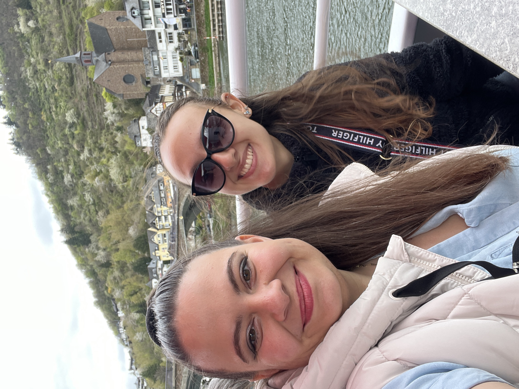 Two women are posing for a selfie on a boat in the city of Cochem, Germany. The woman in the foreground wears a light pink puffer jacket over a light blue shirt. The woman behind her wears sunglasses and a black Tommy Hilfiger lanyard. The background includes buildings and a church steeple. The river is visible behind the women.