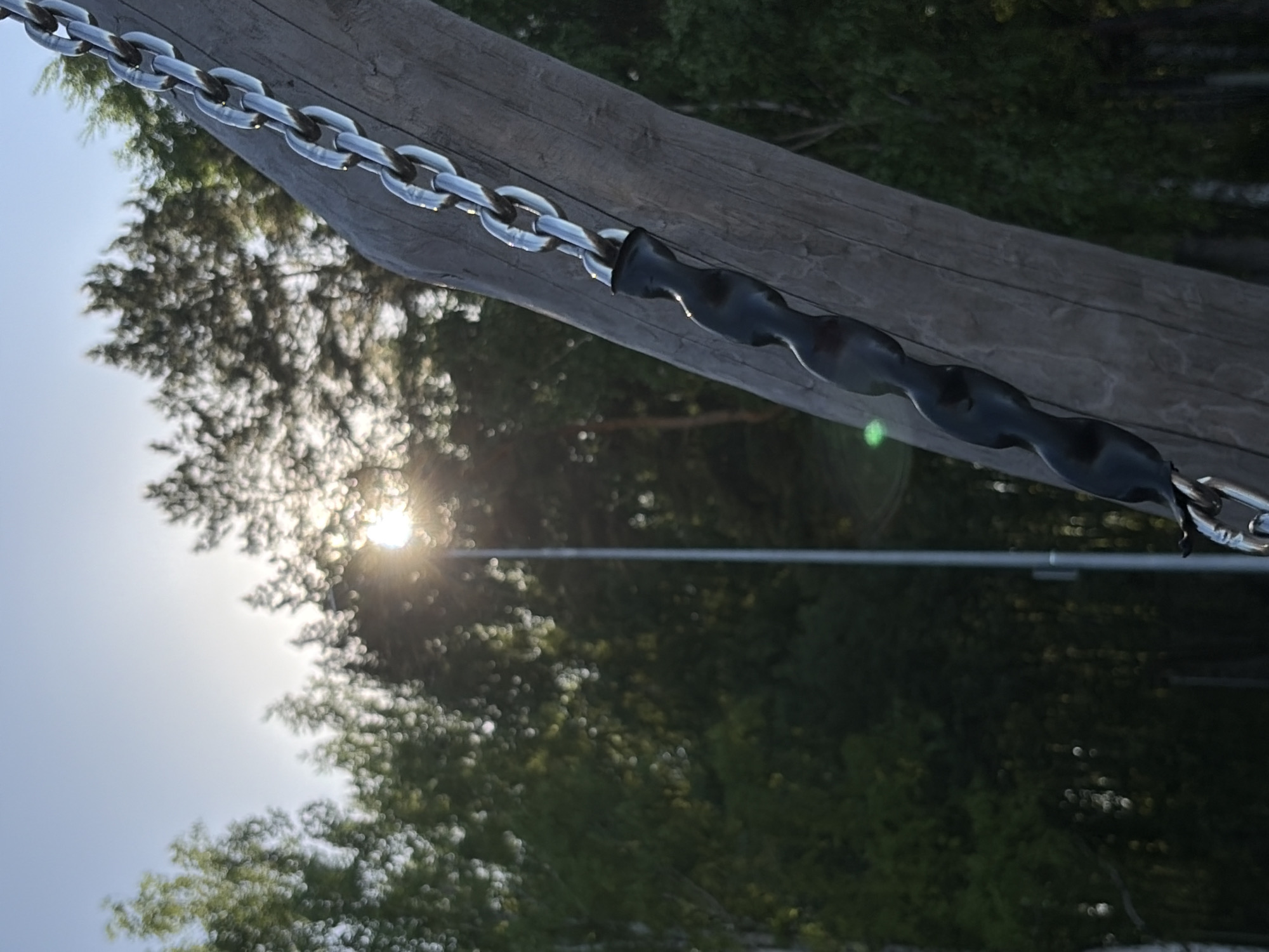 A close-up shot focuses on a swing set chain hanging from a wooden beam. The chain links are made of silver metal, and the section closest to the swing seat is covered in a black, ribbed material. The background reveals a park-like setting, with trees and a bright sun shining through the foliage. A horizontal, thin, metal bar is also visible behind the chain and wooden beam. The location is Unknown, Unknown.