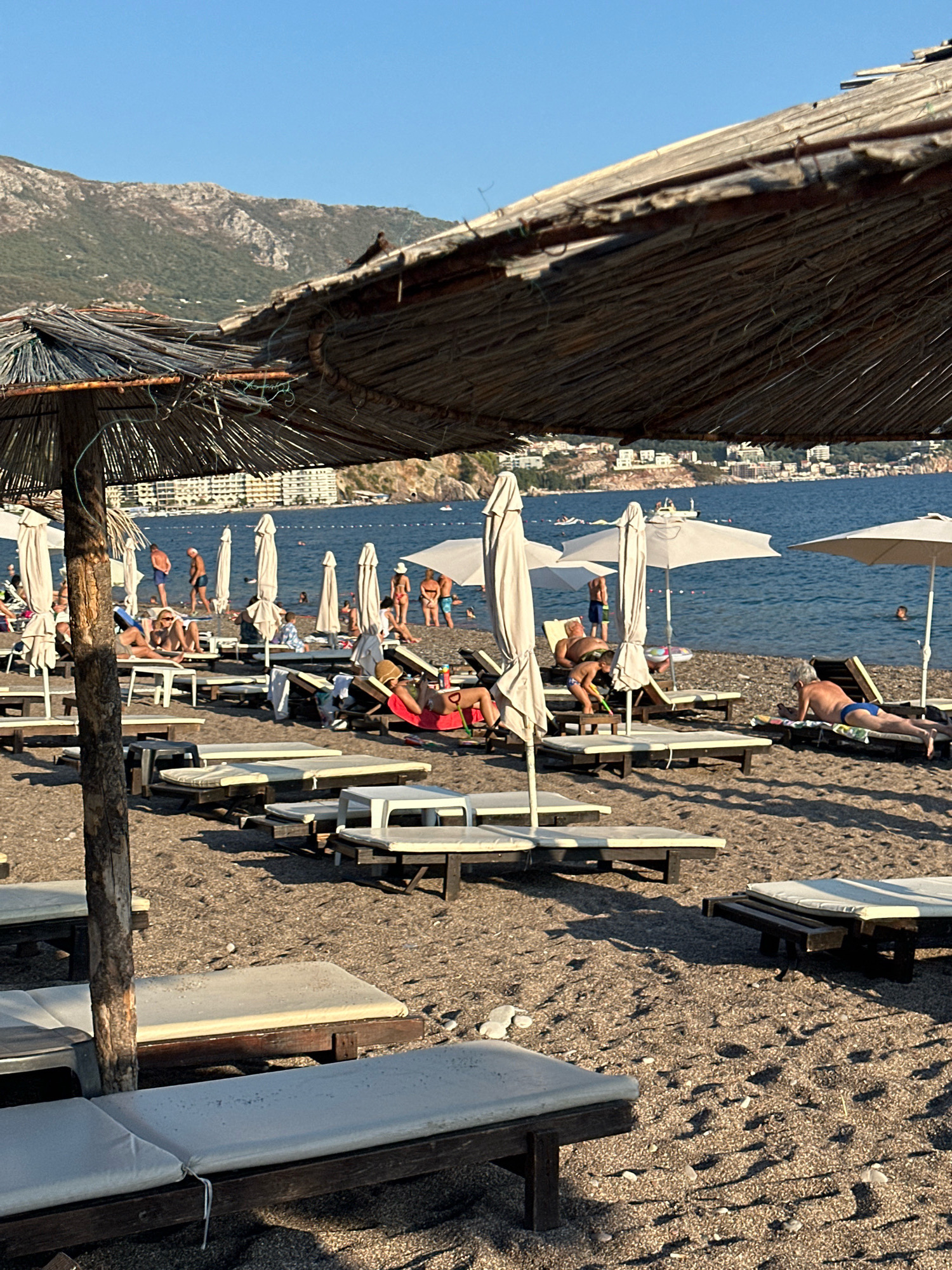 This image depicts a beach scene in Boreti, Montenegro, on a sunny day. The foreground is dominated by several empty and occupied lounge chairs and a large straw umbrella, casting shadows on the sandy beach. The middle ground reveals more beachgoers relaxing on lounge chairs or standing near the water. Several white umbrellas are also visible. In the background, the blue sea stretches out, with a distant coastline featuring buildings and a mountainous landscape. The overall atmosphere suggests a leisure and recreational environment.