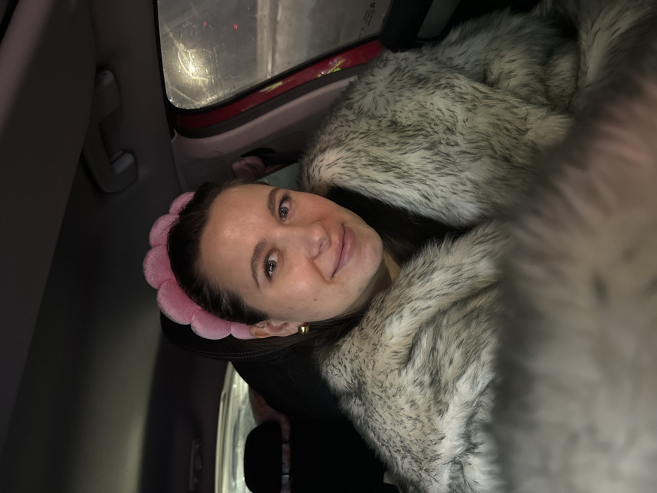 A female subject, appearing to be in her 20s or 30s, is seated within the interior of a vehicle in Akseno-Butyrki, Russia. She has dark hair pulled back and wears a distinctive pink, scalloped headband. Her attire includes a light grey and beige faux fur coat with darker flecks. A gold-colored hoop earring is visible on her right ear. The subject is looking directly at the camera with a faint smile. The car's interior features a dark headliner, an overhead grab handle, and a portion of a car seat headrest behind her. A red vehicle door frame is visible on the right, adjacent to a window appearing smudged or wet. Faint, diffuse lights are visible through the window, suggesting low-light or nighttime conditions outside the vehicle.