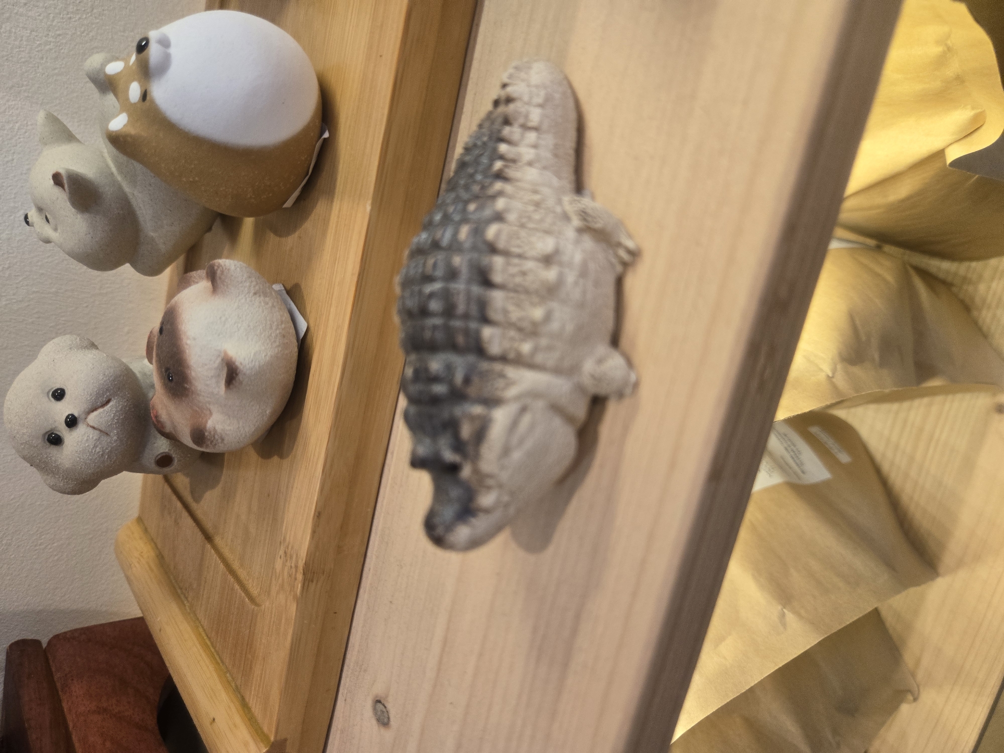 The image shows a wooden shelving unit with various decorative items on display. On the left, there are three ceramic animal figurines, appearing to be a bear, a pig, and a deer or a similar animal. To the right of the animal figurines is a crocodile-shaped object, possibly a ceramic or stone figurine. The background includes a blurry view of what seems to be a shelf filled with brown paper bags. Based on the objects, it is likely taken inside a shop in Budva, Montenegro that specializes in tourist souvenirs or handmade local crafts.