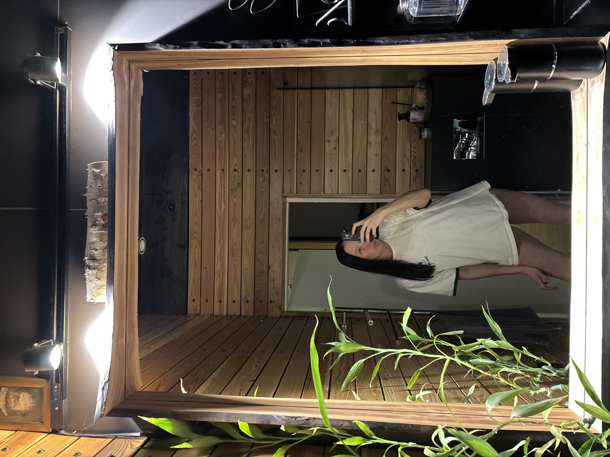 A mirror reflecting a woman taking a picture of herself with her phone is in the foreground. The mirror's frame is made of wood. The woman has long, dark hair and is wearing a white t-shirt. Behind the woman in the mirror is a wall made of light-colored wood planks with dark round accents, and a black wall. There is also a black counter with a faucet. In front of the mirror is a plant with long, green leaves. The location is Chaikovsky, Russia.