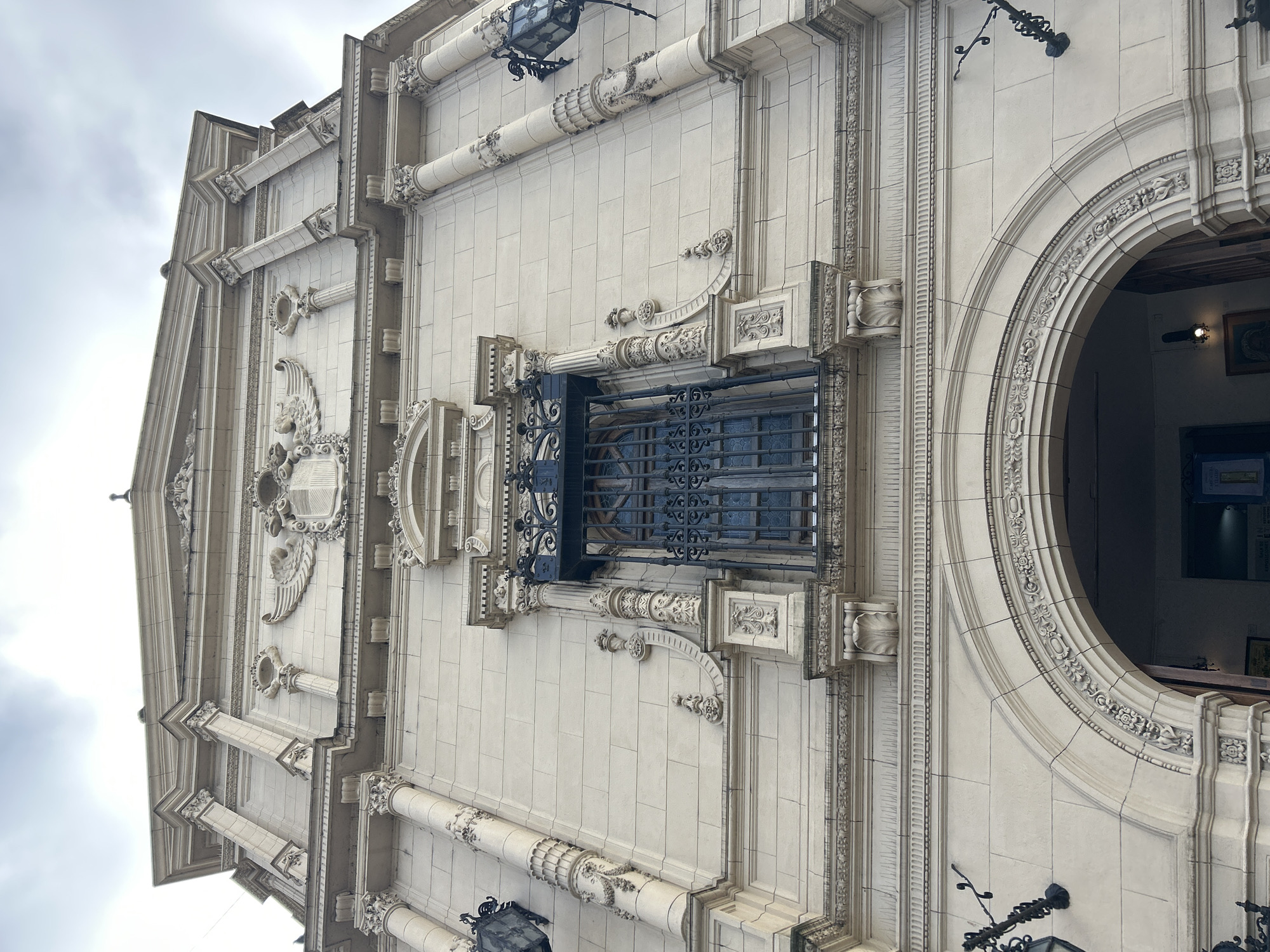 The image shows a low-angle view of the facade of the Teatro Colón in Buenos Aires, Argentina, under a cloudy sky. The building's exterior is made of light-colored stone blocks and features ornate architectural details, including columns, carvings, and a prominent arched entrance. A decorative balcony with a wrought-iron grille is visible above the entrance, with a large window behind it. Black wrought-iron sconces with lamps are attached to the facade on either side. The overall impression is one of grandeur and historical significance.