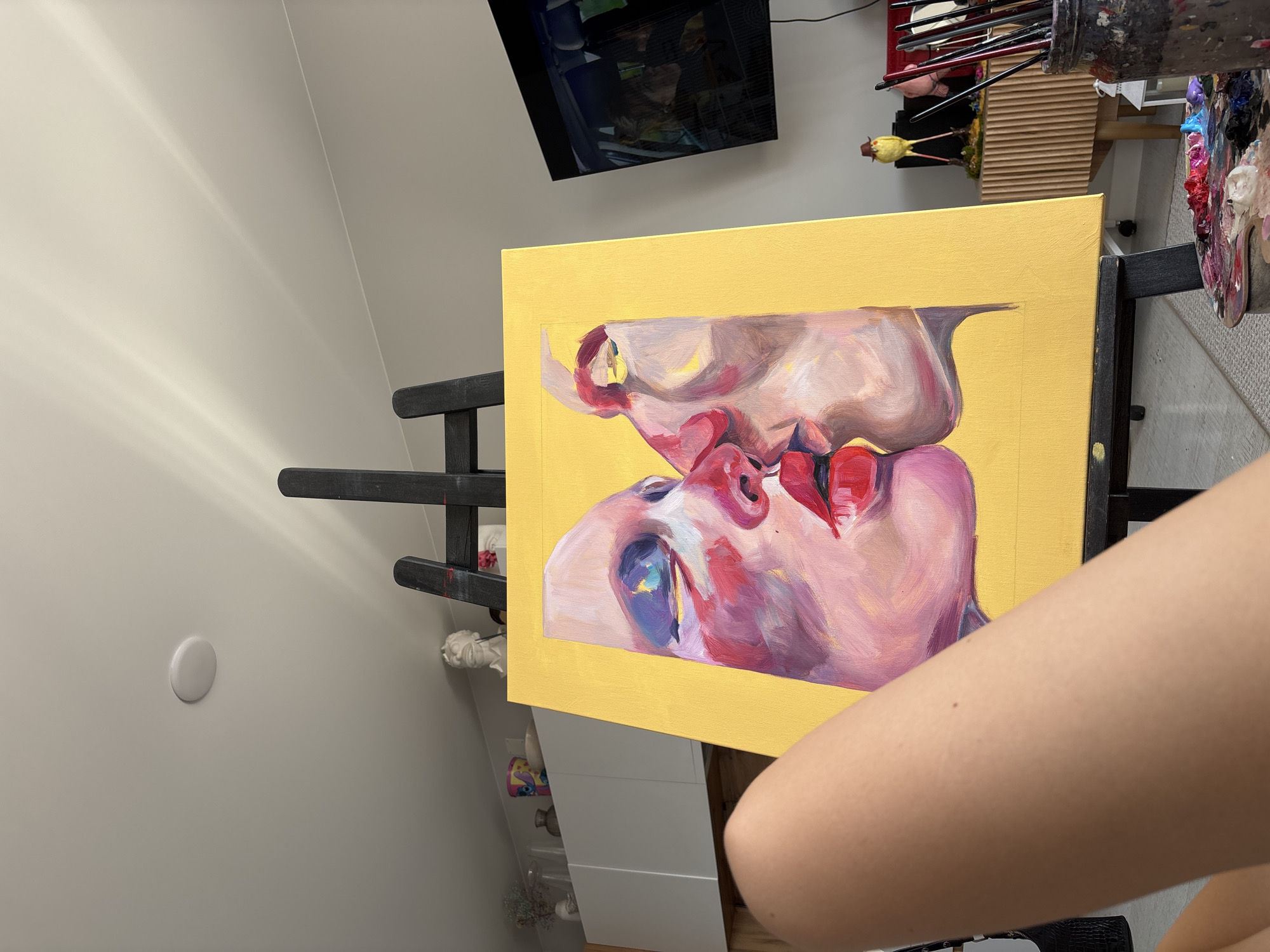 An indoor scene captures an art studio or creative space, prominently featuring a vibrant painting on a canvas held by an easel. The painting depicts two stylized faces in an intimate, close-up embrace or kiss, rendered with expressive, broad brushstrokes in shades of pink, purple, red, and blue, set against a bright yellow background. The upper face is slightly tilted, with prominent red lips almost meeting the lower face's lips. Part of a human forearm and upper arm is visible in the bottom right, suggesting an artist is either actively working on the piece or has just paused.
To the right of the canvas, a jar brimming with paintbrushes and an artist's palette laden with colorful oil or acrylic paints (red, blue, white, black, green, yellow, purple) are seen, indicating an ongoing creative process. In the background, a flat-screen television is mounted on a light-colored wall, displaying what appears to be a cooking show or similar media. Below the TV, a small yellow bird figurine wearing a brown hat is perched on a shelf. The walls are a neutral light tone, with subtle light reflections, suggesting natural daytime illumination. A white circular fixture, possibly a light or smoke detector, is visible on the left wall. The setting implies a dedicated creative environment, possibly in a home or personal studio in Krasnoyarsk, Russia. No visible text is present in the image.