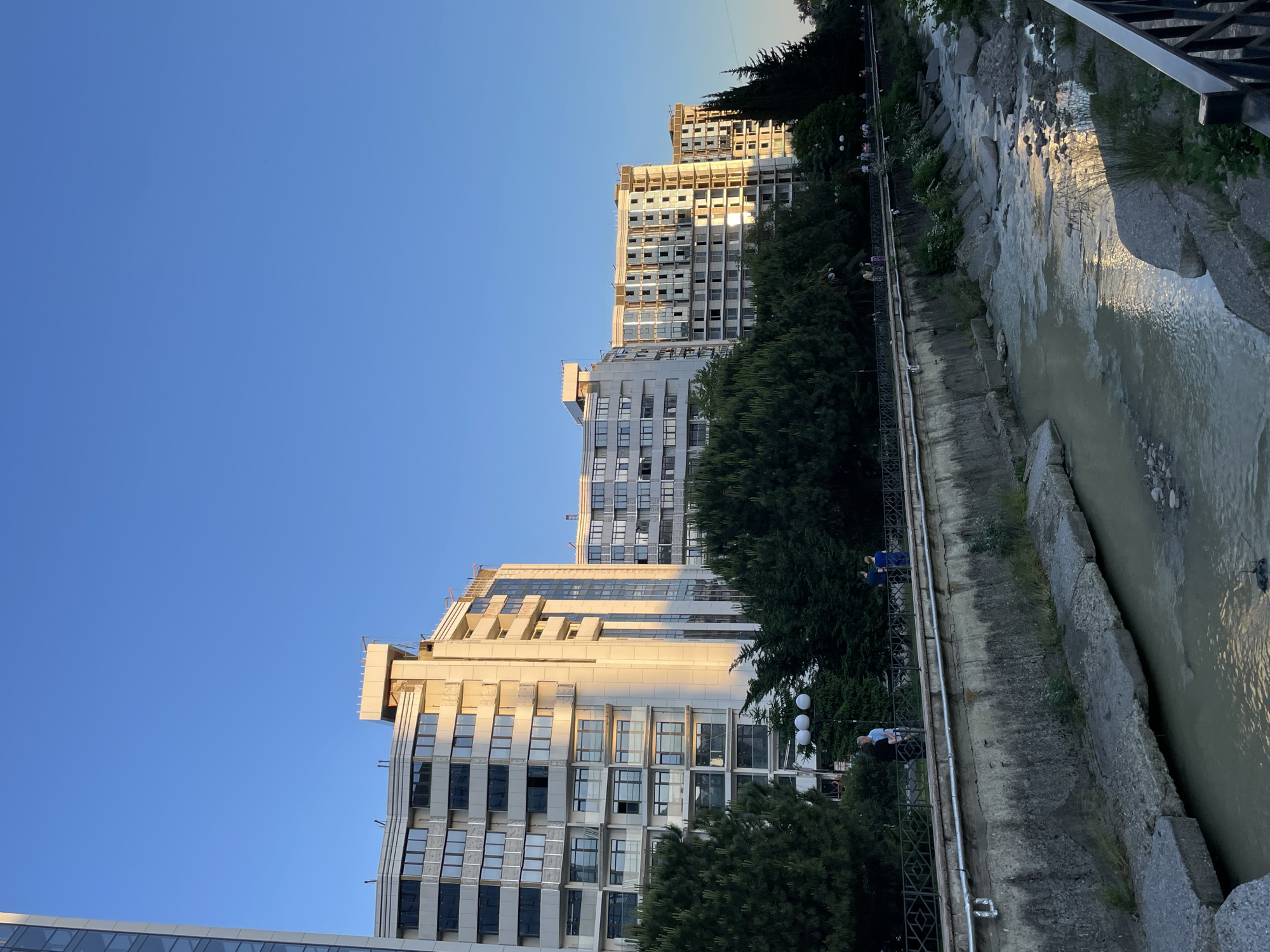 The image captures an urban scene in Sochi, Russia, under a clear, bright blue sky during late afternoon or early evening, evidenced by the warm, golden light illuminating one side of the structures and casting shadows.
In the midground and background, a series of modern, multi-story buildings with numerous windows line a street. The facades of these buildings, particularly those on the right and center, are brightly lit by direct sunlight, appearing in shades of white and gold. Several of these structures show signs of ongoing construction or recent completion, with exposed upper levels, scaffolding-like elements, or incomplete window sections. A taller building is partially visible on the far left.
In the foreground, a narrow river or canal flows from right to left, contained within a man-made channel comprising weathered concrete retaining walls and some natural rock formations. The water appears turbid or murky brown, with reflections of the golden sunlight visible on its surface in various patches.
Above the concrete river wall, a paved pathway or promenade is present, delineated by a dark metal ornamental railing. This pathway is lined with a dense arrangement of mature trees, including tall conifers and broader green deciduous trees. Several individuals and small groups of people are visible on this pathway, some standing near the railing on the left, and others walking further down the path on the right. Streetlights with three spherical lamps are interspersed among the trees. A dark metal fence or railing extends into the lower right corner of the frame.