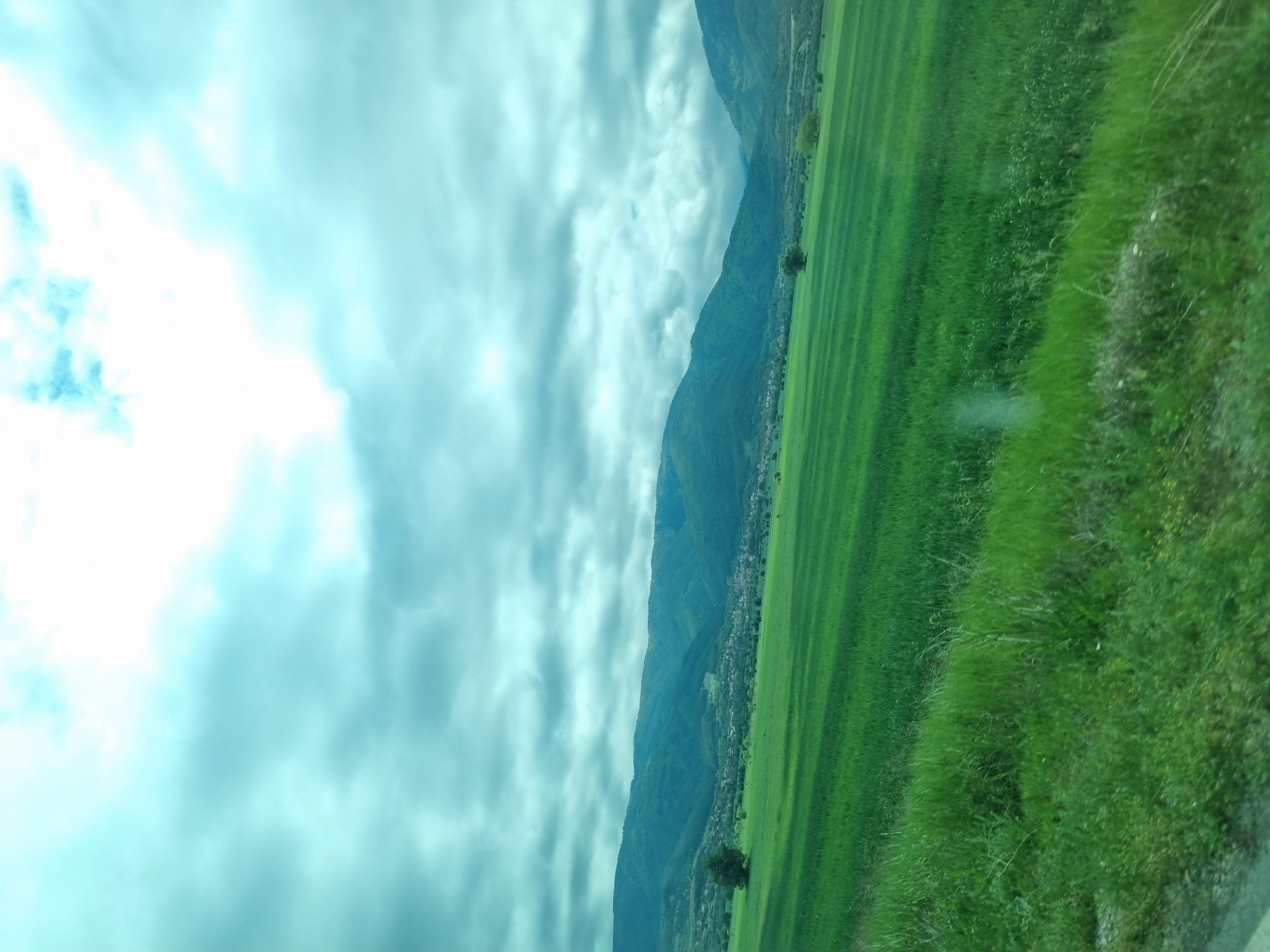 The image shows a landscape featuring a green field and mountains under a cloudy sky. There is a small town at the base of the mountains. The landscape is likely located in or near Yahinovo, Bulgaria, based on the prompt. There are no people visible in the image.
