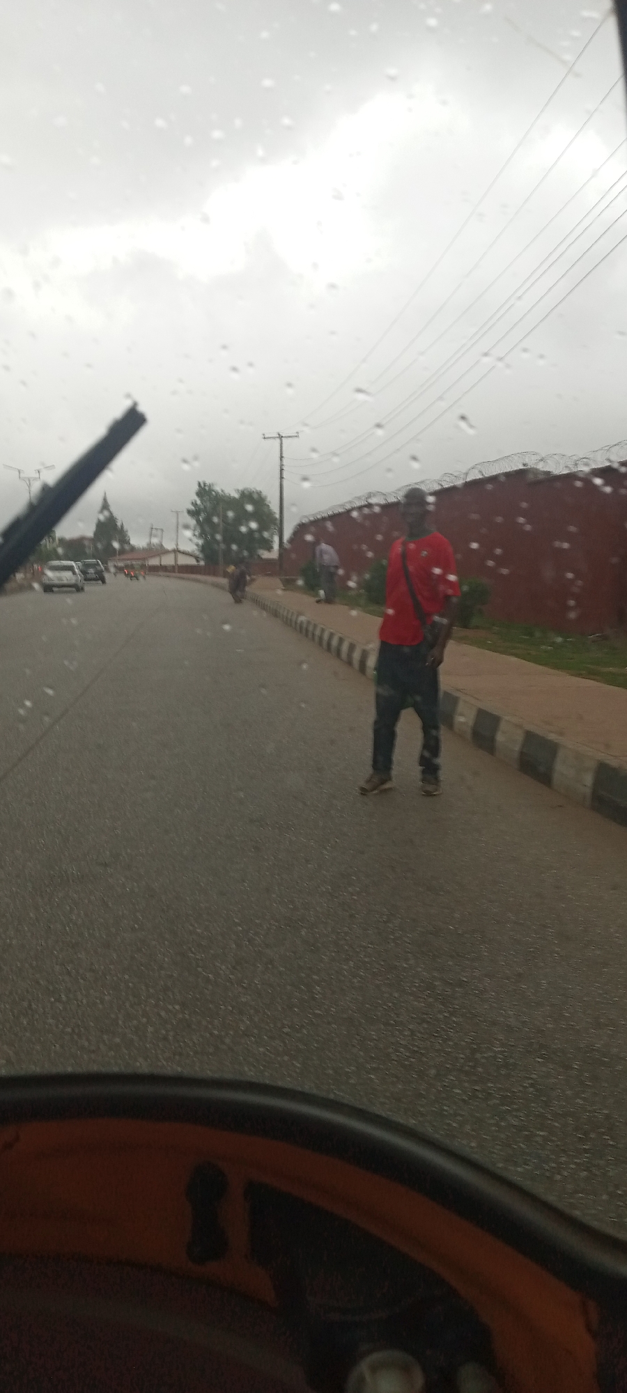 The image, taken from inside a moving vehicle, possibly an auto-rickshaw based on the visible part of the frame, shows a man standing in the middle of a paved road on a cloudy, rainy day in what is likely Jos, Nigeria, given the details present. The windscreen is covered with raindrops, and the wiper is visible on the left side of the frame. The man is wearing a red shirt, dark pants, and has a dark bag slung over his shoulder. He stands on the asphalt road with what appears to be a concrete curb and narrow strip of green vegetation alongside him. Beyond this, a red brick wall topped with barbed wire is visible, partially obscured by trees. In the background, there are other vehicles on the road and a few pedestrians. Utility poles with power lines are visible in the sky. The road surface is wet and reflects the overcast sky.