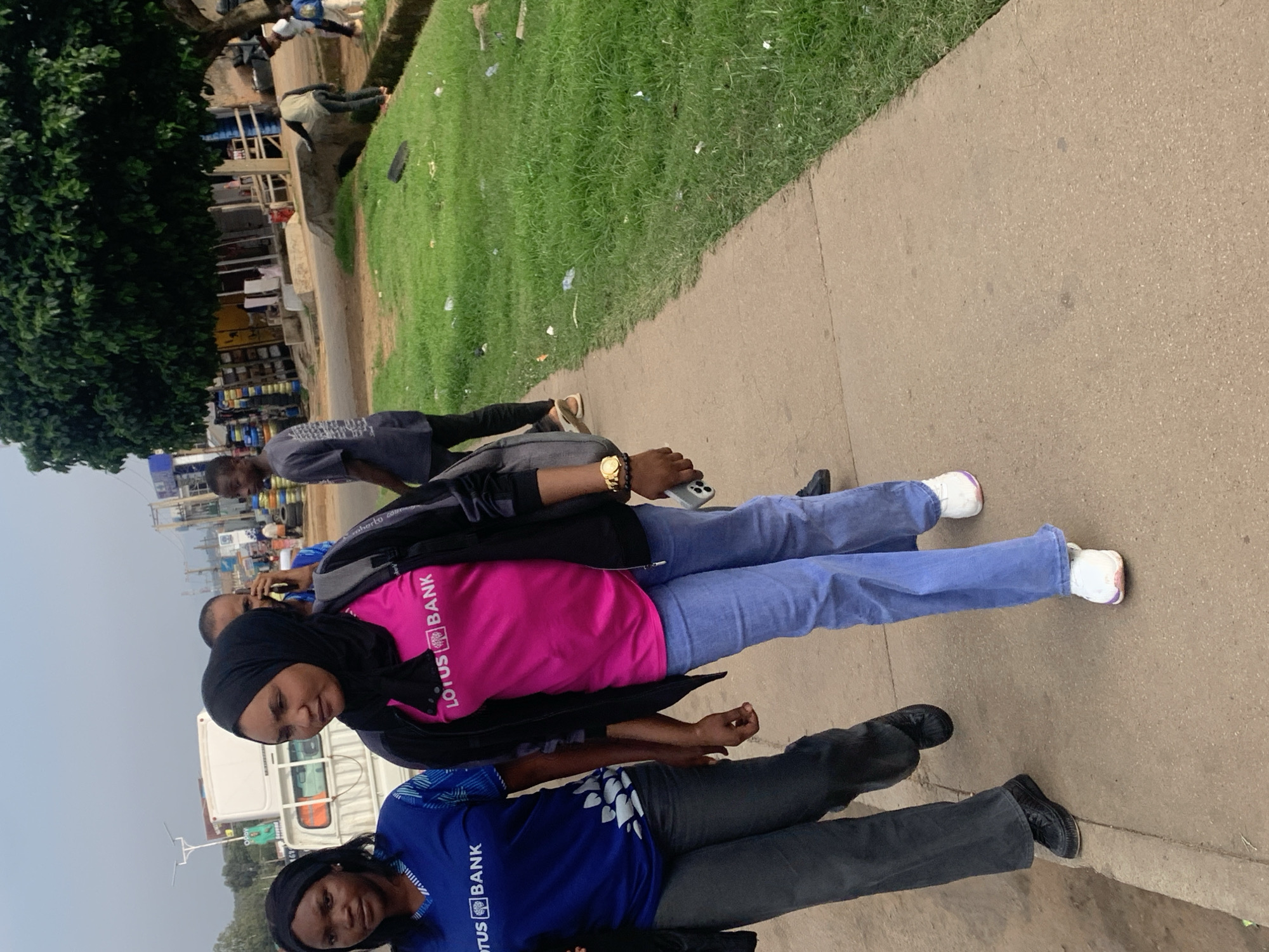 The image shows a group of people in an outdoor setting in Gura Top, Nigeria. Two women are prominently featured in the foreground, both wearing t-shirts with "LOTUS BANK" printed on them, along with a stylized lotus flower logo. One woman wears a pink t-shirt and a black hijab, while the other wears a blue t-shirt and a black headband. They are walking on a paved surface, likely a sidewalk, with a grassy area to their left.
Behind them, other individuals can be seen, including a person wearing a grey t-shirt and another in a black jacket. There's a white truck visible in the background, suggesting a street or roadside environment. The overall scene appears to be daytime, with bright lighting. The people's expressions are neutral. The presence of the bank's branding on their attire indicates they might be employees or participants in an event related to Lotus Bank. The setting suggests an urban or semi-urban environment in Nigeria.