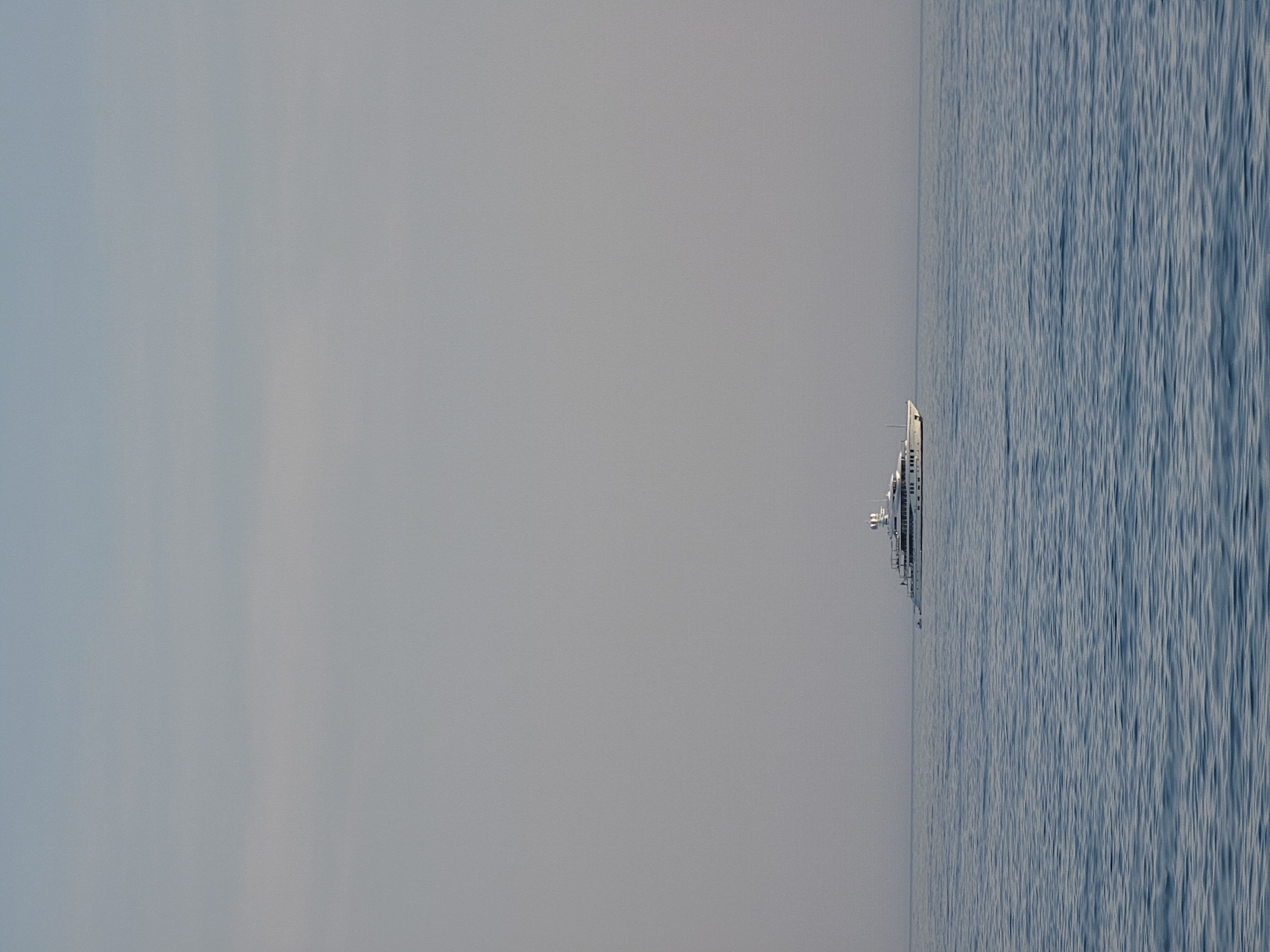 This media file captures a serene seascape with a large, white yacht sailing on calm, blue waters. The yacht is positioned in the right half of the frame, oriented vertically, suggesting the image has been rotated. The vessel appears to be modern, with multiple decks and windows visible. To the left of the yacht, the sky transitions from a pale, hazy grey near the horizon to a lighter, almost white hue at the top of the frame. The water exhibits a textured surface with subtle ripples, reflecting the muted light of what appears to be a cloudy day, possibly late afternoon or early morning given the soft illumination. The horizon line is sharp and distinct, separating the sea from the sky. There is no discernible text within the image. The scene evokes a sense of tranquility and vastness, with the solitary yacht emphasizing the expansive, open ocean. The location context of Quarteira, Portugal, suggests this could be a view off the coast of the Algarve region.