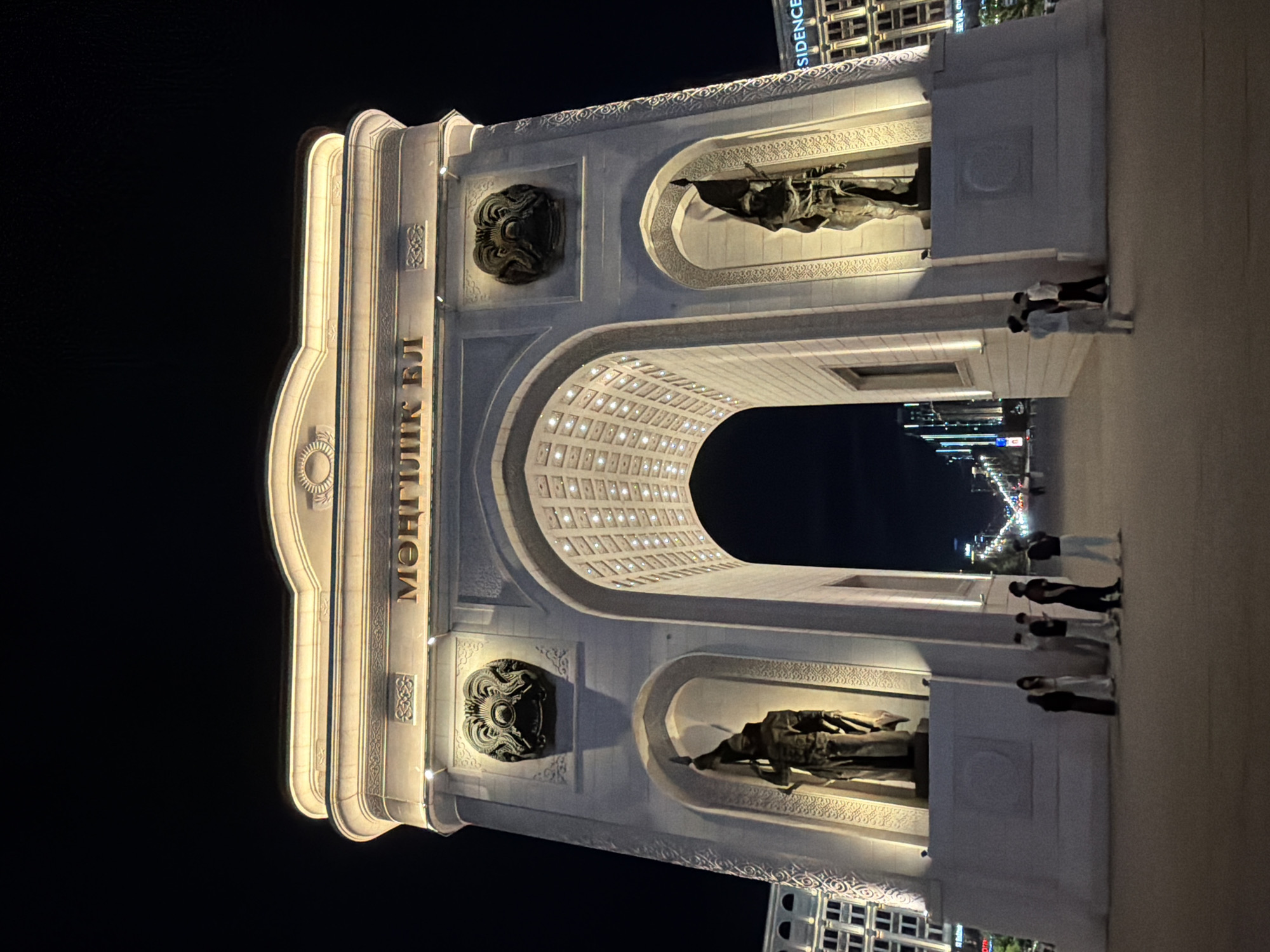 The image depicts the Monument of Independence in Astana, Kazakhstan, at night. The monument, illuminated with golden lights, features a large archway with intricate carvings and two statues of warriors standing in niches. On the arch's facade, there are inscriptions in Cyrillic and circular emblems. In the foreground, a group of approximately six individuals, dressed in casual attire, are gathered on a paved plaza. Behind the monument, a street lined with buildings and illuminated with streetlights is visible, extending into the distance. The overall scene captures a nocturnal urban landscape centered around a prominent historical monument.
