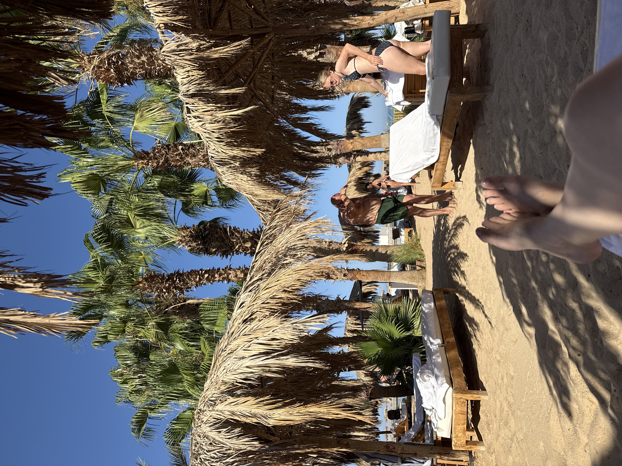 This image depicts a beach scene in Hurghada, Egypt, characterized by palm trees, thatched umbrellas, and lounge chairs on a sandy shore. In the foreground, a person's feet are visible, resting on a lounge chair. Two people are standing near lounge chairs in the middle ground; one, a man wearing green shorts, is facing away from the camera, while a woman in a black polka-dot bikini and sunglasses is facing the camera, her hands resting on a lounge chair. Two other figures in swimwear are seen in the background, near the water. The environment is sunny with a clear blue sky and dappled shade cast by palm fronds and umbrellas.