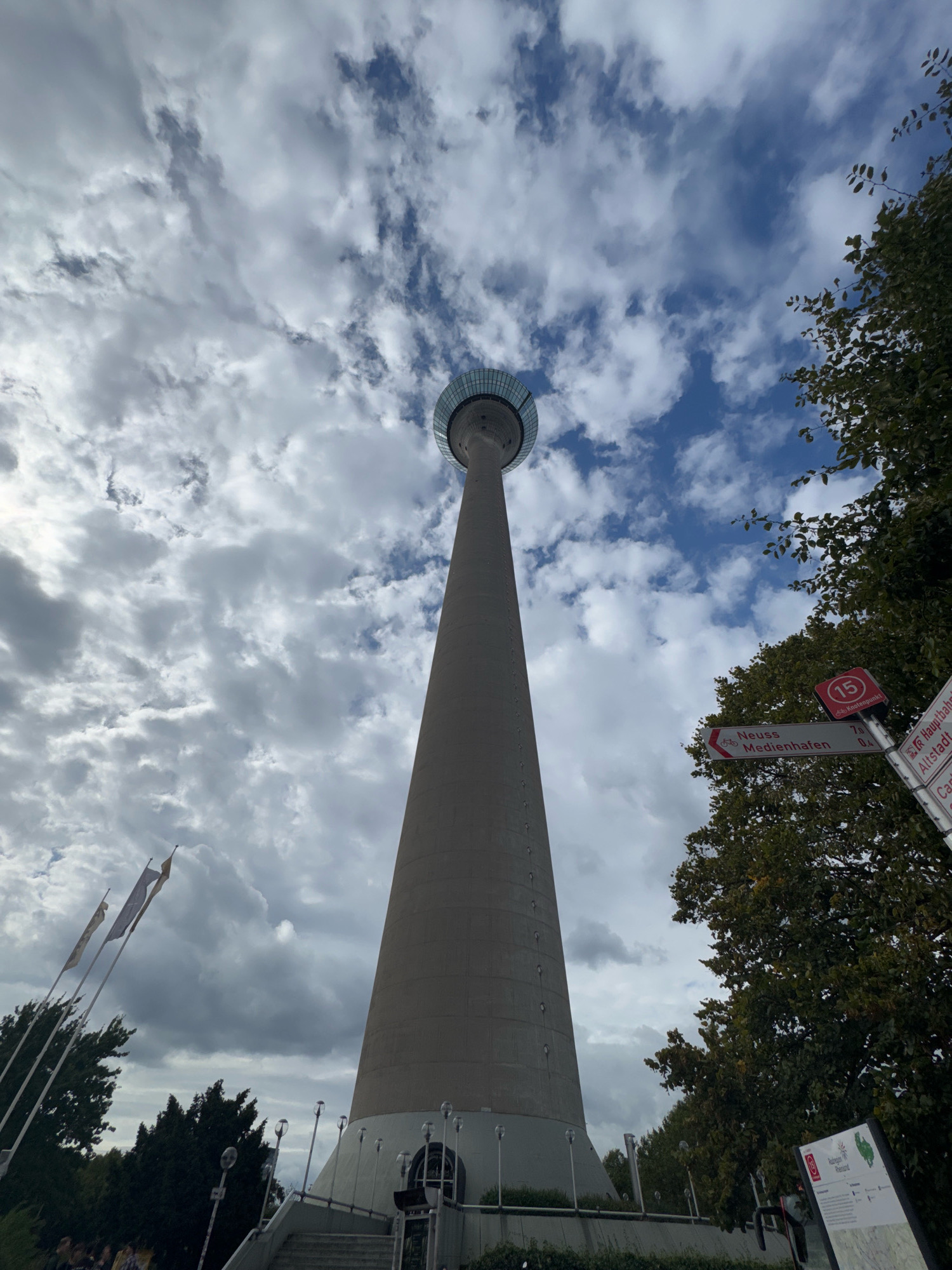 The image shows the Rheinturm, a telecommunications tower located in Düsseldorf, Germany, on a cloudy day. The tower is a tall, concrete structure with a circular observation deck at the top. In the foreground, there are trees, flags, and street signs pointing to various locations including "Neuss Medienhafen" and "Hauptbahnhof". A few people are visible near the base of the tower. The sky is filled with dramatic clouds, with patches of blue sky showing through.