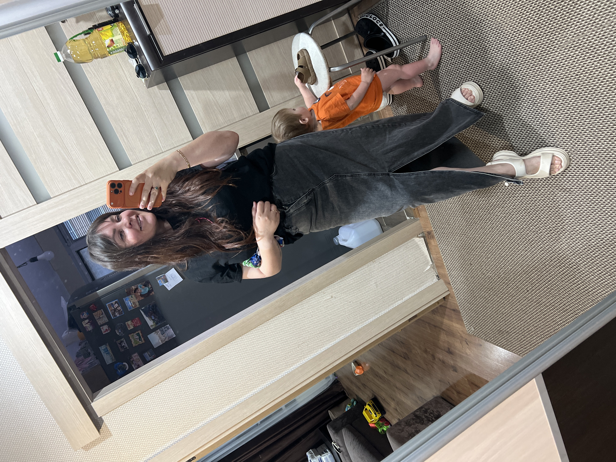 A woman in a black t-shirt and a long grey denim skirt with a high slit takes a mirror selfie in a room. She is wearing white platform sandals. Next to her, a toddler in an orange t-shirt and diaper stands reaching for a brown sandal on a small white stool. The floor is covered with a patterned rug and hardwood. In the background, through a doorway, a kitchen with a refrigerator is visible, adorned with magnets. A bottle of cooking oil and sunglasses sit on a shelf in the reflection of the mirror.