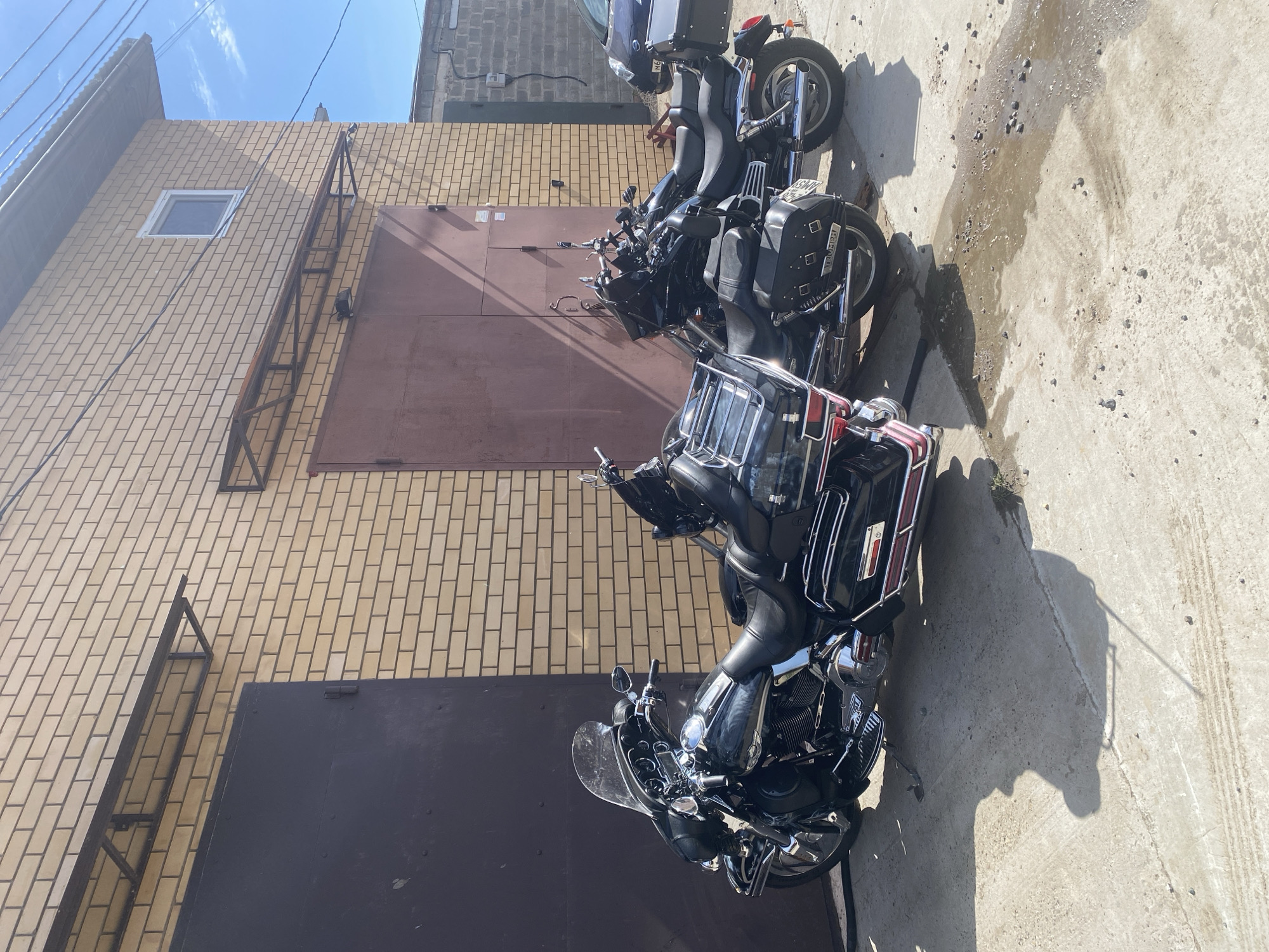 The image shows three motorcycles parked on a concrete surface adjacent to a multi-story brick building. The building has a brown door and a ladder affixed to the side. The sky is visible in the upper portion of the image. Based on the location metadata, the image was taken in the city of Chaikovsky, Russia.