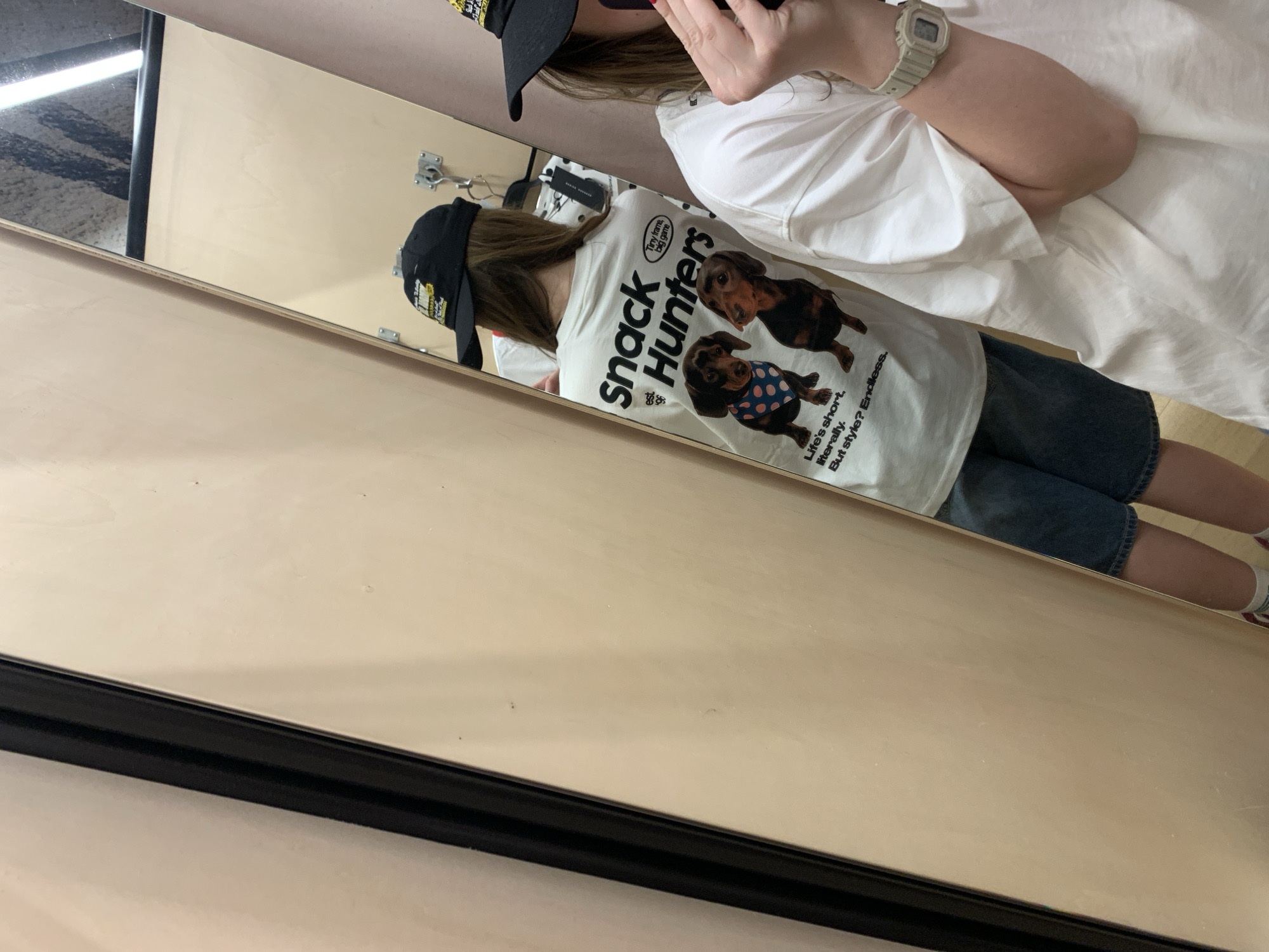 An angled mirror selfie captures a person indoors, likely in Belgrade, Serbia, reflecting their outfit. The person, seen mostly from the back in the reflection, has long brown hair and wears a black baseball cap with yellow detailing, partially visible text that reads "Snack Hunters". They are dressed in a light-colored (off-white or cream) short-sleeved t-shirt and blue denim shorts.
The t-shirt features prominent black text on the back, reading "Snack Hunters". Below this, there are two graphic illustrations of dachshund dogs. One dog is positioned above the other, while the lower dog wears a blue bandana with white polka dots. Smaller text on the shirt includes phrases like "Tiny frame, big game", "Life's short. literally.", and "But style? Endless." The person also wears a light grey digital wristwatch on their left wrist.
The immediate surroundings visible in the reflection include a plain, light-colored wooden or laminate wall with two metal hooks mounted on it. In the upper left corner of the mirror, a reflection of a patterned dark and light rug or carpet can be seen, along with a bright linear light source. The mirror itself has a thin black frame along its bottom edge. The overall image is taken at an oblique angle, with the mirror occupying a significant portion of the frame.