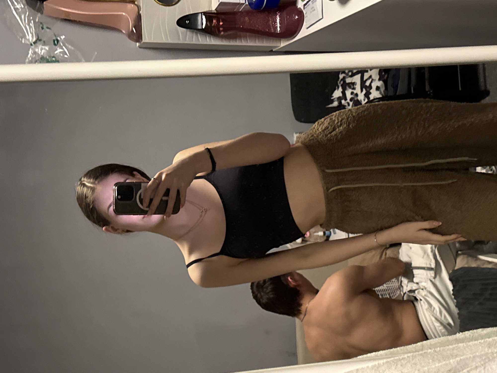 A woman is taking a selfie in a mirror. She is wearing a black top and brown sweatpants. A shirtless man is lying in a bed next to her, wearing light-colored shorts. The reflection shows a cluttered shelf above the mirror. The location cannot be determined from the image.