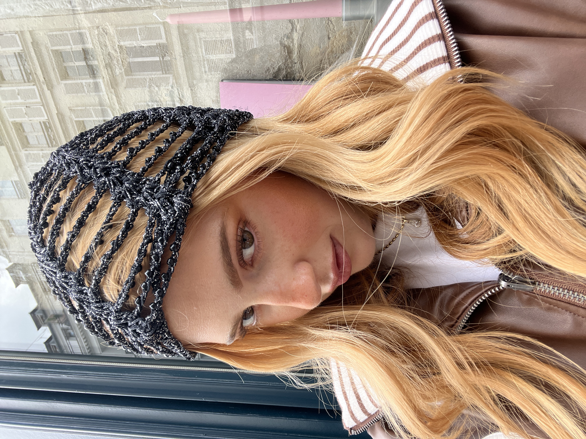 A young blonde woman is seen in a close-up portrait, framed by a black, beaded hair covering. She gazes downward, her gaze slightly left. The background, visible through a window, reveals a Parisian cityscape. The architecture is characterized by classic stone buildings with repetitive window patterns, typical of Paris. The woman is wearing a brown leather jacket with a white and brown striped lining peeking out. A pink object, possibly a notebook or cosmetic item, is partially visible behind her head. The composition is dominated by the woman's face and hair, set against the backdrop of Paris.