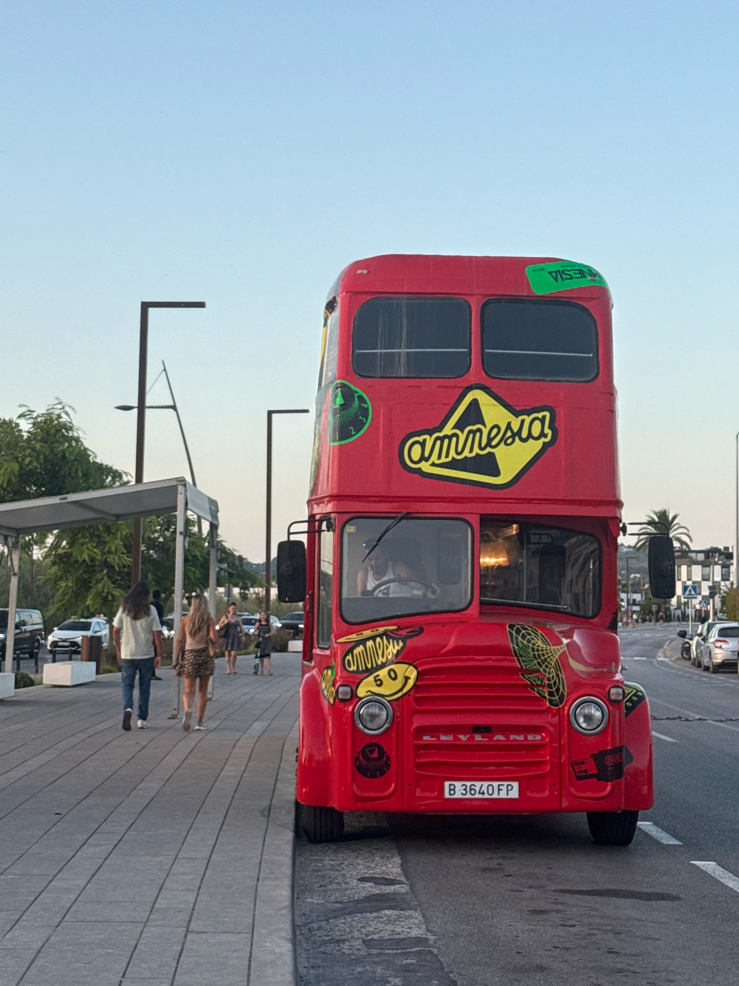 This is a full-frame, eye-level, medium shot of a red double-decker bus parked on a paved street in Ibiza, Spain. The bus, likely a former public transport vehicle, is decorated with several stickers, most notably a large yellow and black sticker on the side of the upper deck that reads "amnesia". A green sticker on the top right of the upper deck also reads "amnesia". The license plate on the front of the bus reads "B 3640 FP".
In the background, several people are walking on a sidewalk. Two individuals, a man with long hair and a woman in a leopard-print skirt, are walking away from the camera. Behind them, other people are visible, as well as parked cars and trees. The sky is clear and a light blue, suggesting it is either early morning or late evening. The overall scene suggests a tourist or entertainment district.