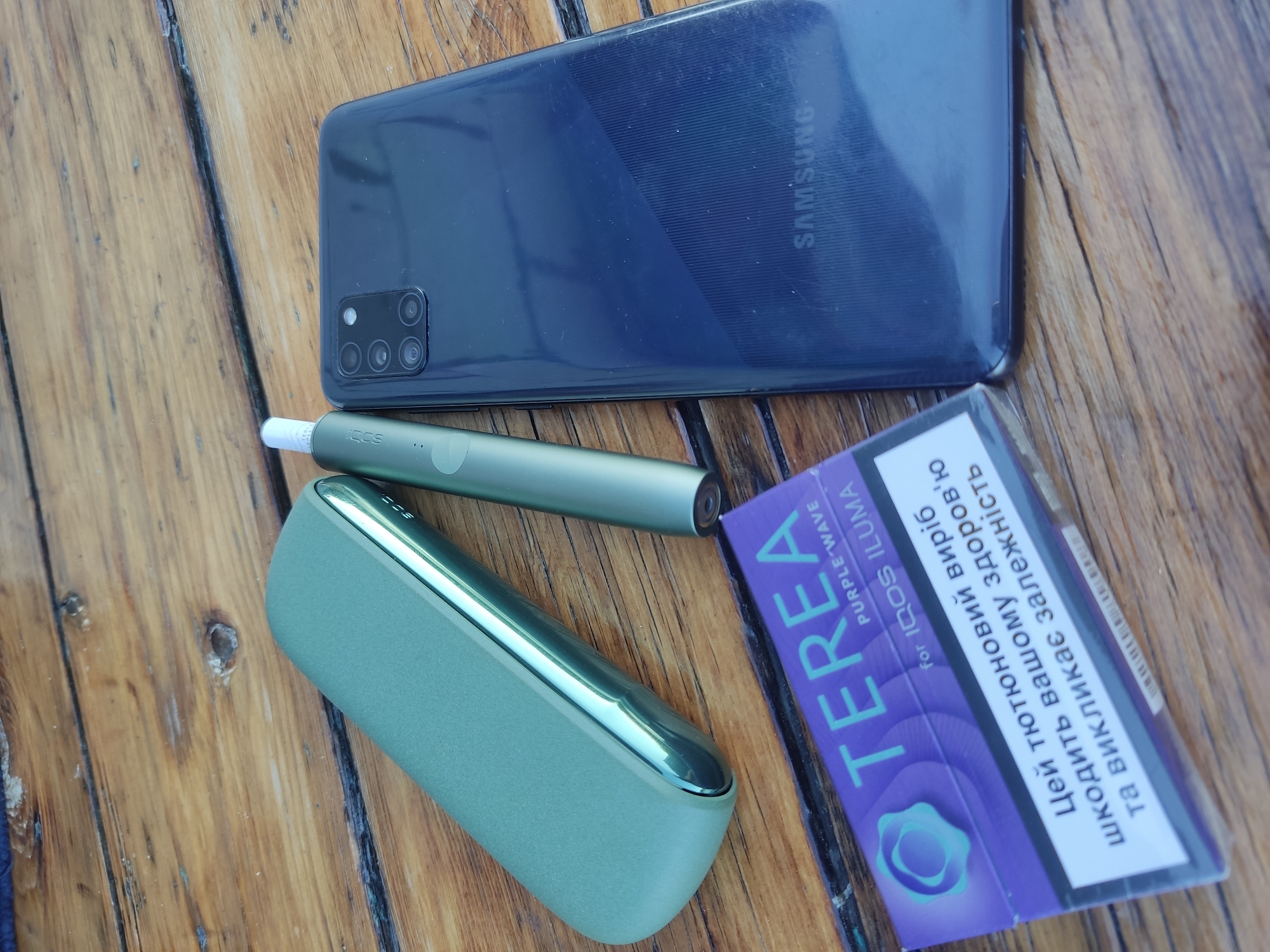 A Samsung smartphone and a green IQOS device with its holder and a pack of Terea Purple Wave for IQOS Iluma cigarettes are placed on a wooden surface. The scene is shot from a top-down perspective. There is no indication of the location in the city of Radisne, Ukraine.