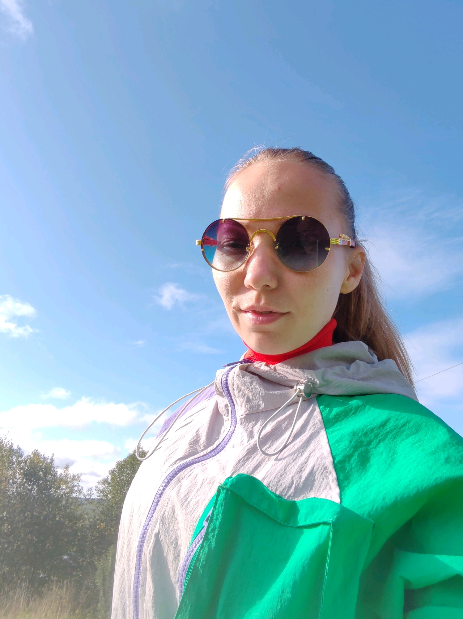 A young woman is posing for a selfie outdoors, with a clear blue sky and some wispy clouds in the background. She is wearing large, round sunglasses with gold frames and a red turtleneck. Her jacket is a color-blocked design, featuring light gray, white, and bright green panels. The jacket has a zipper and drawstring details. A few trees and distant foliage are visible in the lower left portion of the frame. The image is taken from a low angle, looking up at the woman. The setting does not contain any identifiable landmarks or features specific to Чухлэм, Russia.