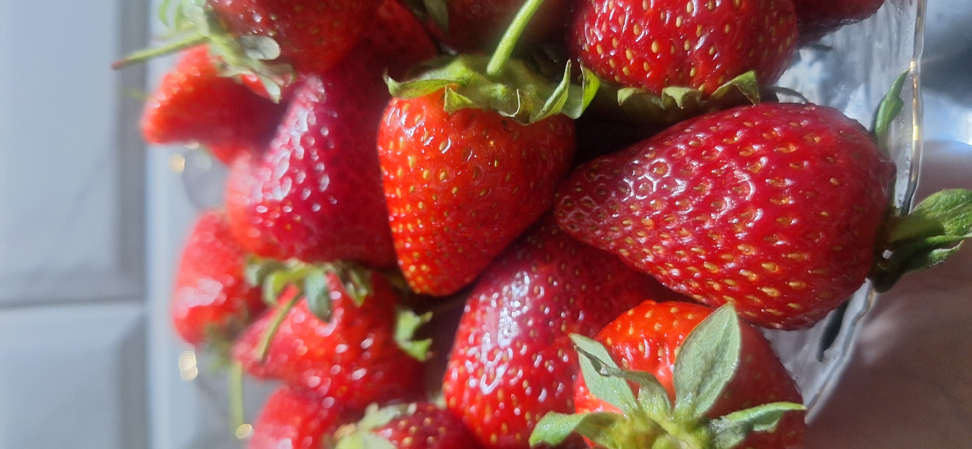 A close-up photograph shows a cluster of fresh, red strawberries with green stems, seemingly contained within a clear glass jar or container. The strawberries are brightly lit and fill the frame, obscuring most of the background. The background shows what appears to be a white tiled wall. There is no information in the image that indicates the location is Moscow, Russia. No people are visible.