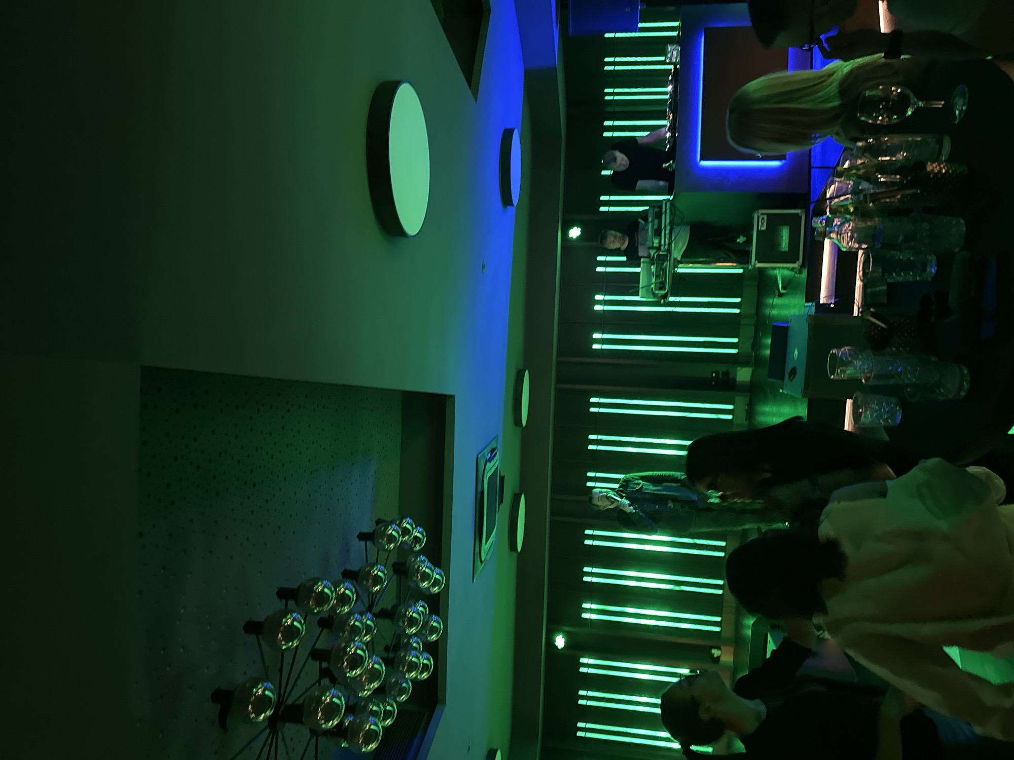 The image shows the interior of a bar or club in Kazanlak, Bulgaria. The lighting is predominantly green and blue, created by neon lights on the wall and circular light fixtures. There are several people present, some gathered at a bar with glasses and bottles on it, while others stand nearby. One person is likely operating a DJ or sound system. The wall has a patterned design with a decorative light fixture consisting of multiple bulbs.