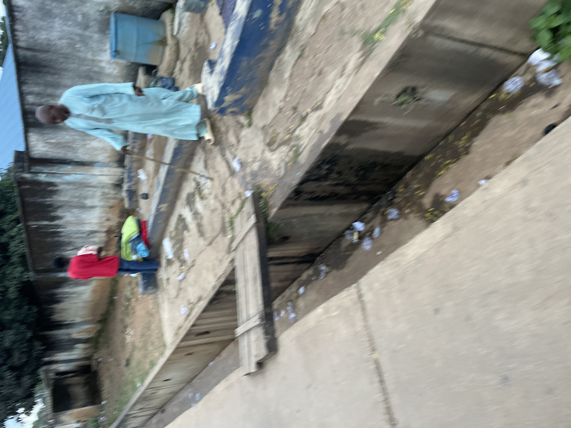 In the city of Jos, Nigeria, a scene unfolds outdoors in what appears to be a semi-urban or transitional area. A drainage ditch runs prominently through the foreground, filled with murky water and scattered debris, including plastic waste and leaves. A makeshift wooden plank bridge spans the ditch in the mid-ground, allowing passage across it.
On the right side of the ditch, an older man, likely a local resident, is walking with a wooden stick for support. He is wearing a traditional light blue, long-sleeved robe and sandals. He appears to be moving away from the viewer, towards the background.
On the left side, nearer to the background, a younger person, dressed in a red t-shirt and blue jeans, is standing and handling some items. Next to them, a pile of colorful folded fabrics, including a bright green one, is stacked on what might be luggage or bags. The background shows a weathered wall, a building with an open doorway, and dense foliage from trees overhead, suggesting a location that is not densely built up. The overall impression is one of a utilitarian outdoor space, perhaps a service area or a less-maintained pathway in Jos.
