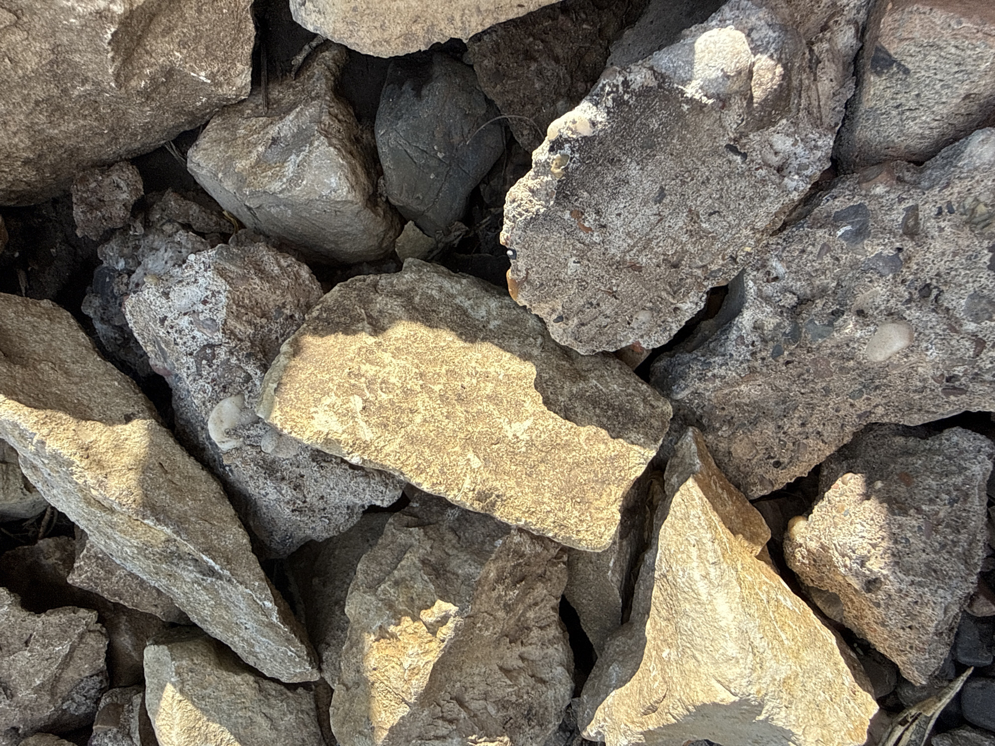 The image depicts a collection of rocks and concrete rubble. The stones vary in size, shape, and texture, with some showing signs of weathering and fragmentation. The lighting is uneven, creating shadows and highlights across the surfaces of the rocks. The overall setting appears to be outdoors, possibly an urban or construction site in an unknown location. No people, animals, or other distinct objects are visible. The primary action is the accumulation of these materials together. There is no apparent interaction beyond their physical proximity.