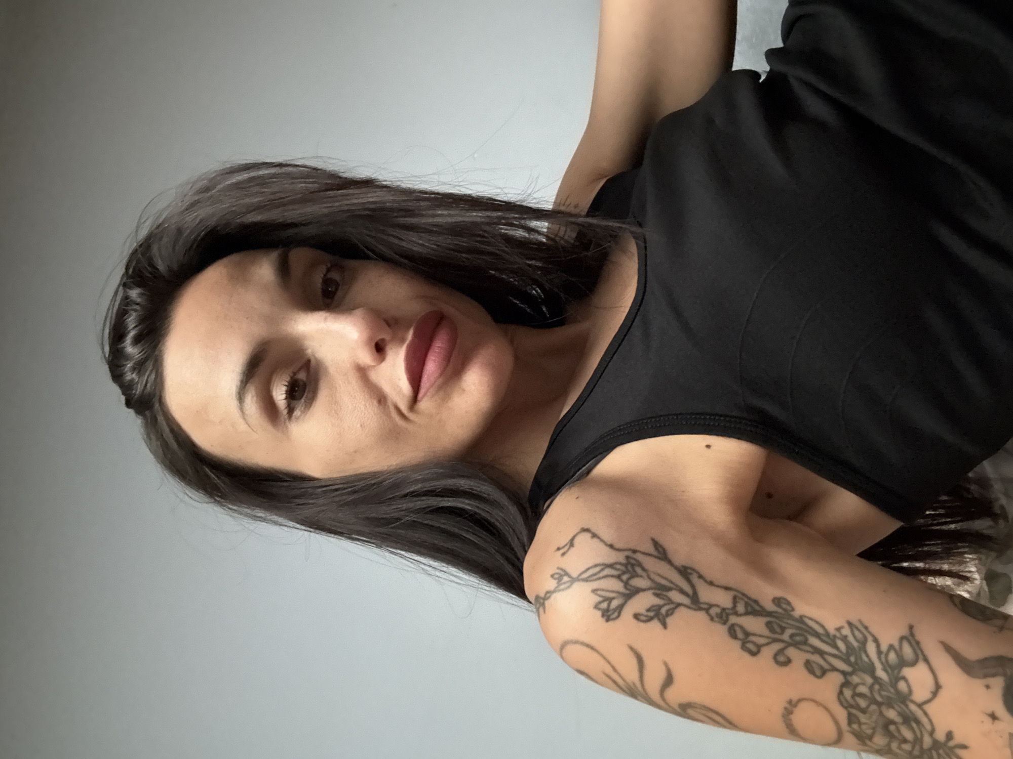 A woman with long, dark hair is wearing a black tank top and has a floral tattoo on her upper arm. The background is a plain, light gray wall. The image appears to be taken from a slightly low angle. No specific location in Pleven, Bulgaria is identifiable.