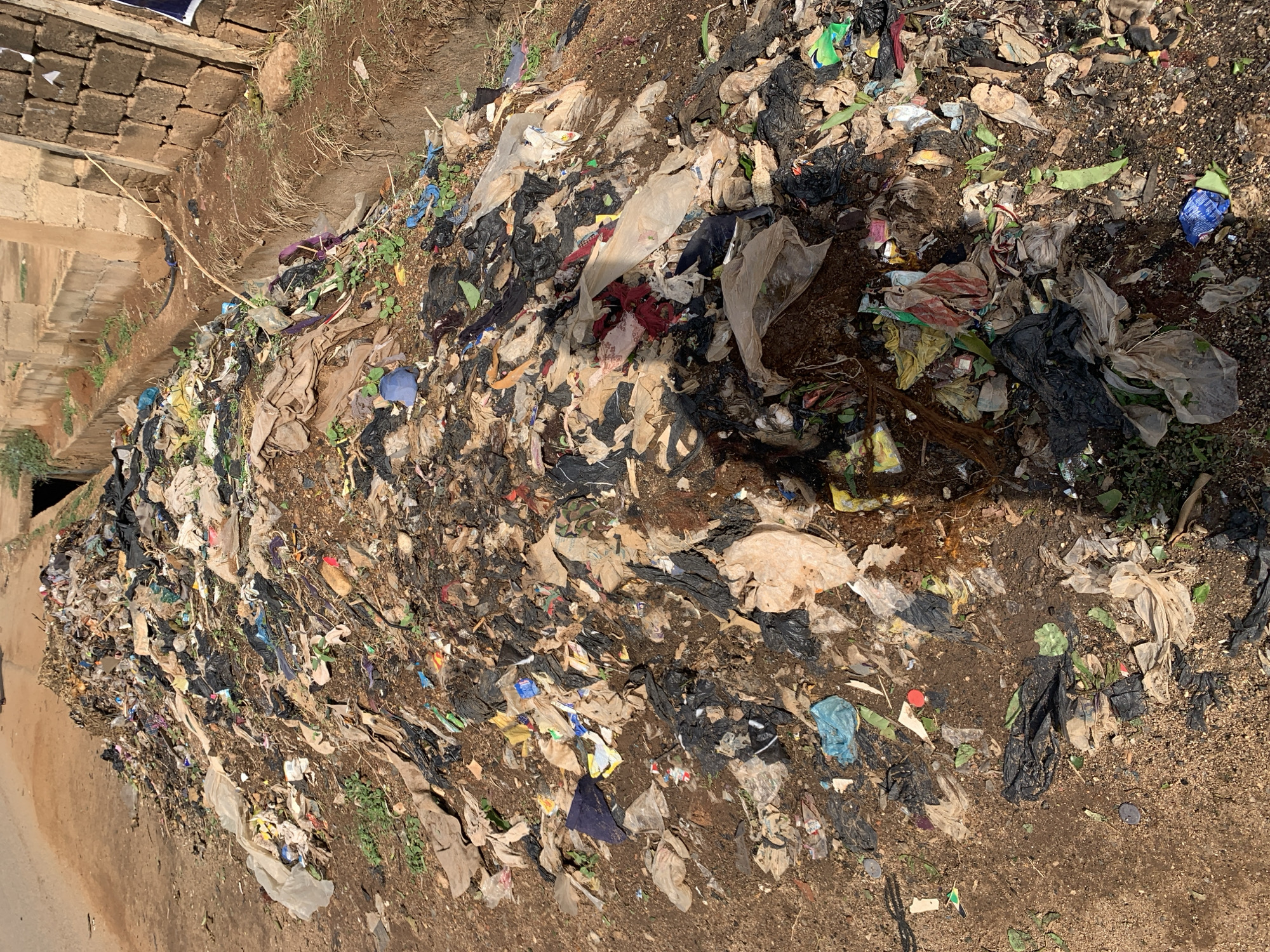 The image depicts a large accumulation of discarded waste, primarily plastic bags and other refuse, dumped along the side of a road or pathway adjacent to a drainage ditch. The location is in the city of New Stadium, Nigeria. The waste is scattered across the ground, mixing with the soil and some sparse vegetation. The ditch is lined with concrete blocks. There is no evidence of people, animals, or movement in the scene.