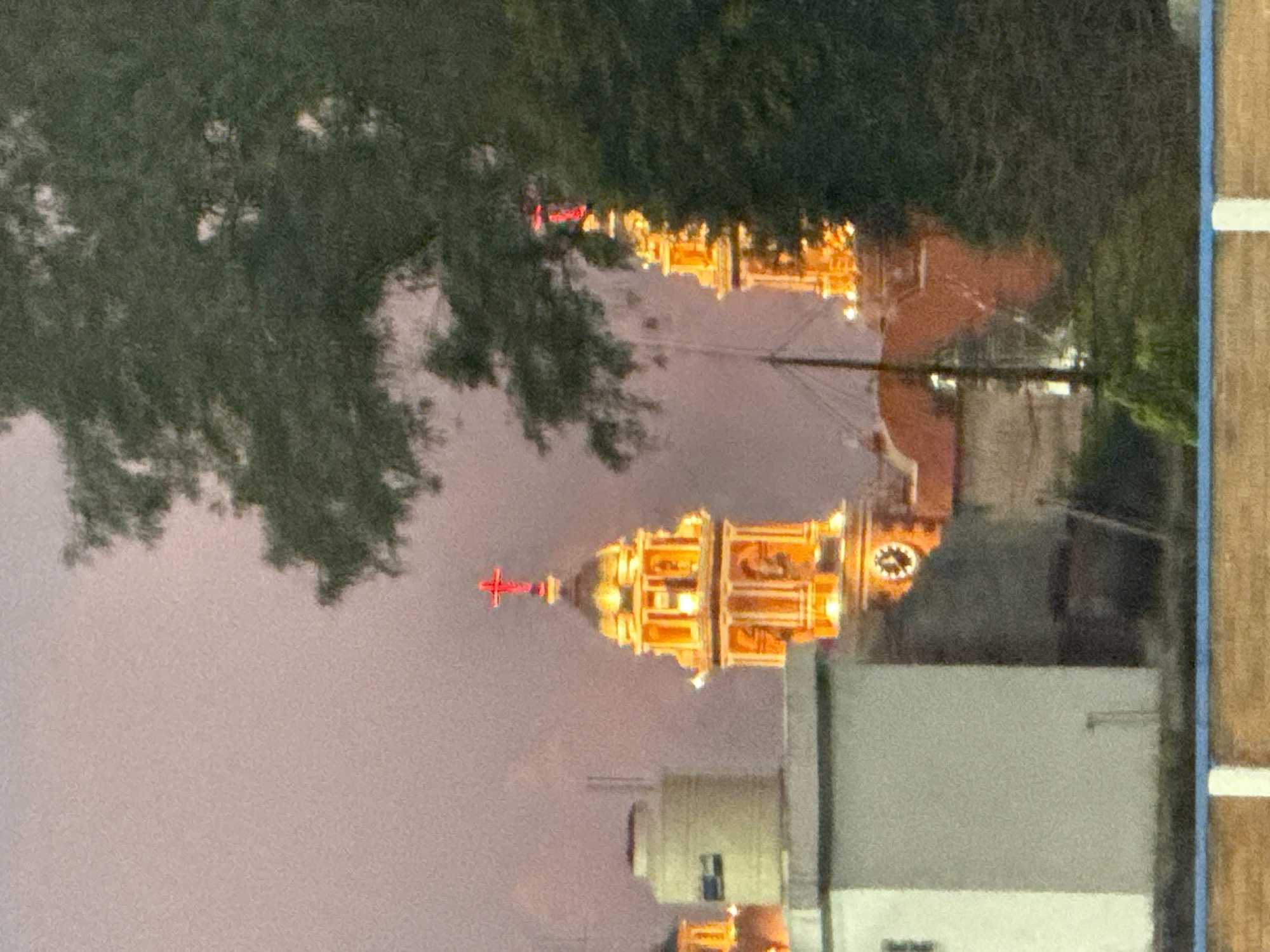 The image captures a nighttime view of the illuminated towers of the Santuario de la Virgen de los Remedios church in San Andrés Cholula, Mexico. The church is partially obscured by trees in the foreground, and the sky is a dusky pink. A residential building is visible on the right side of the frame. The image appears to have been taken from an elevated position.
